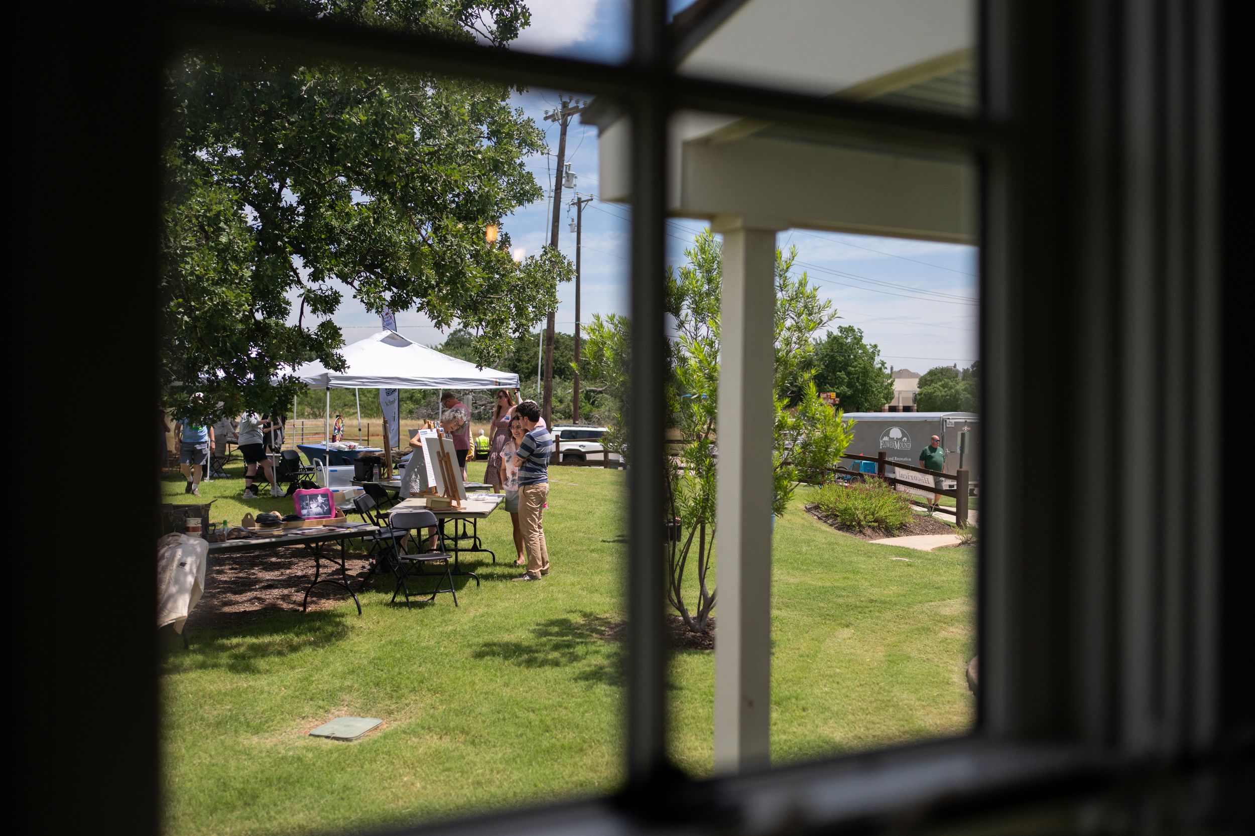 View through a window of an outdoor gathering with people, tables, and a white tent on a sunny green lawn.