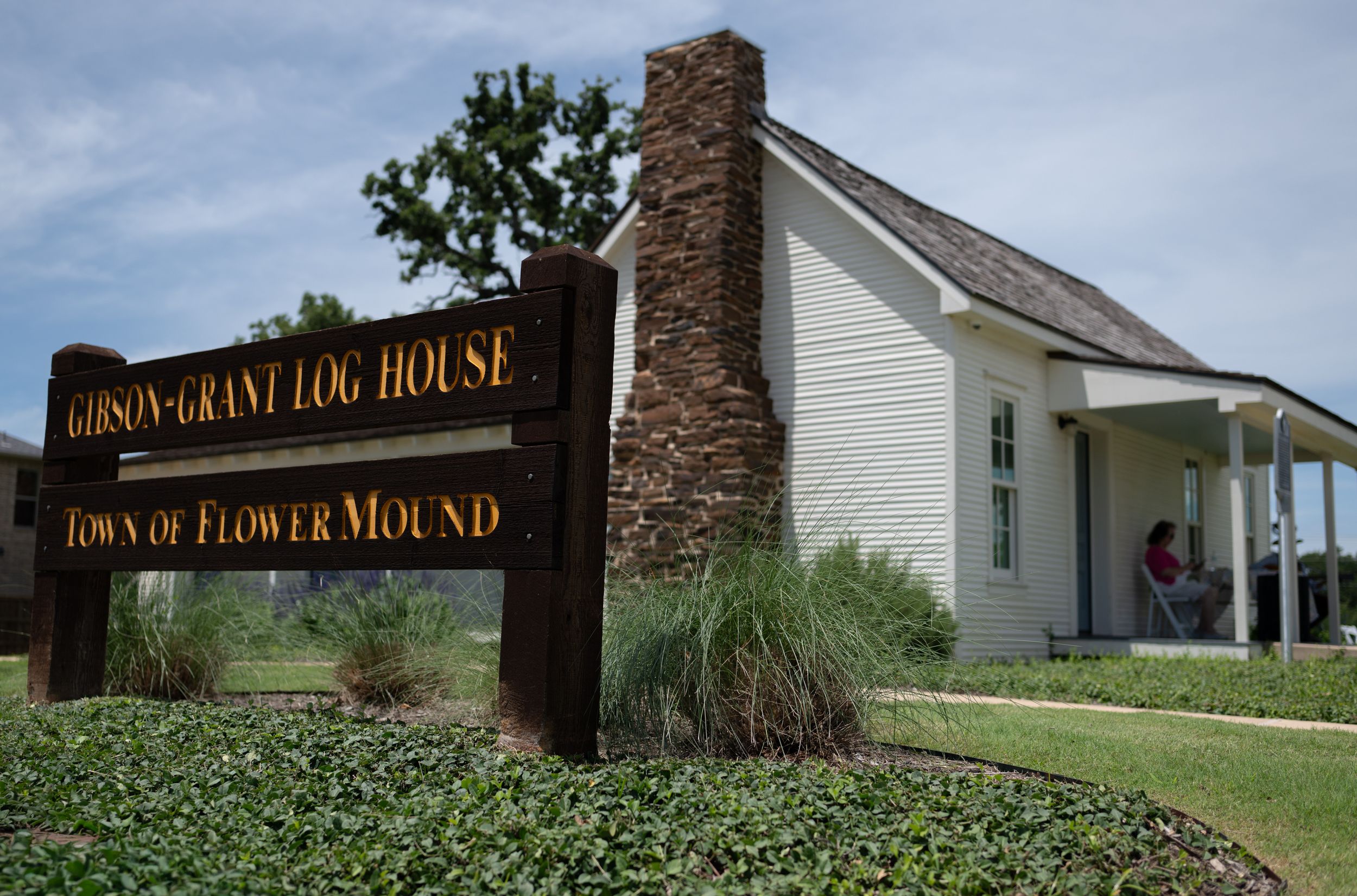 Gibson-Grant Log House historic landmark sign in Flower Mound, Texas, with white wooden building in background.