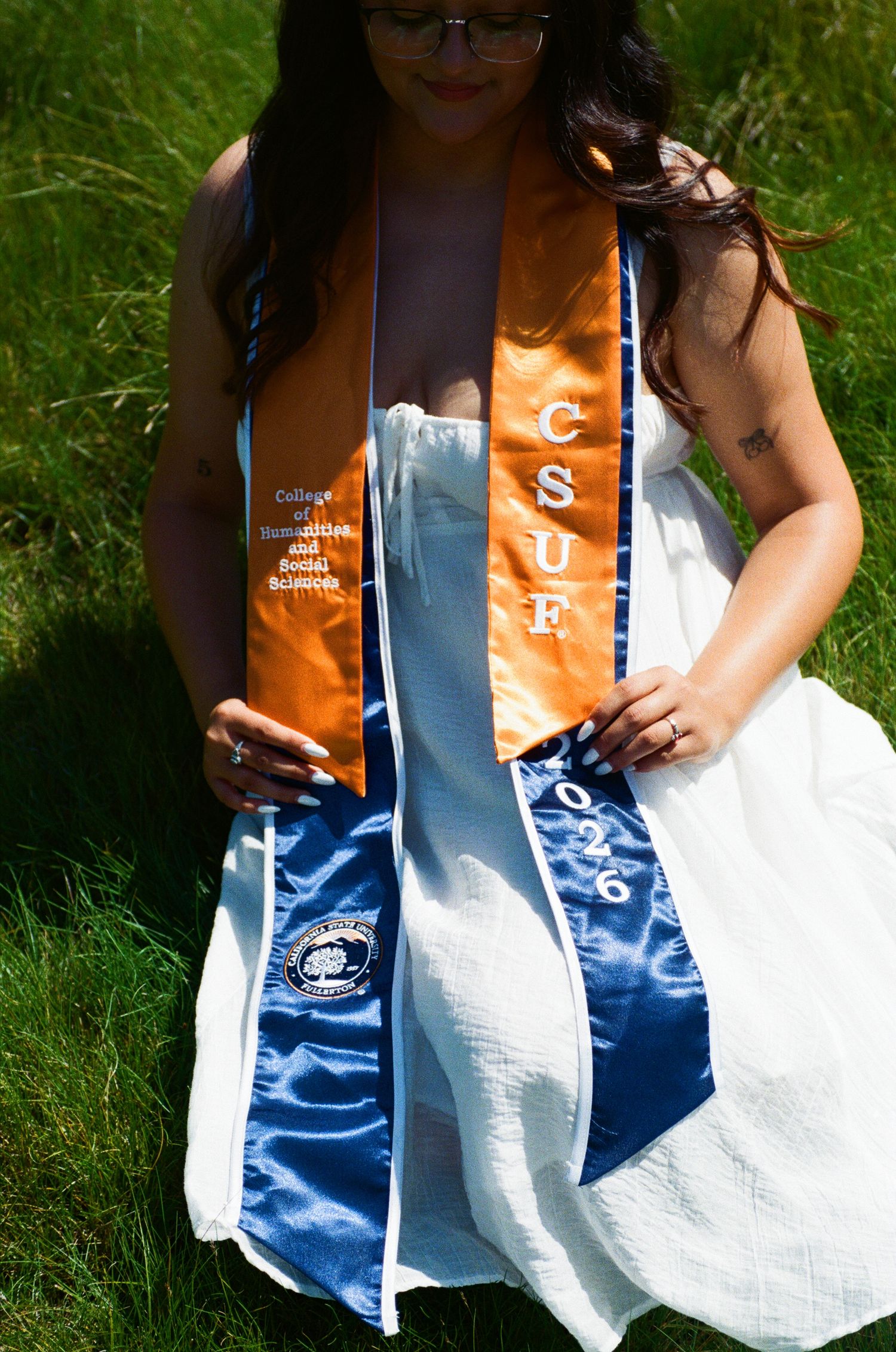 CSUF graduate poses outdoors in white dress wearing orange and blue graduation stoles on green grass.