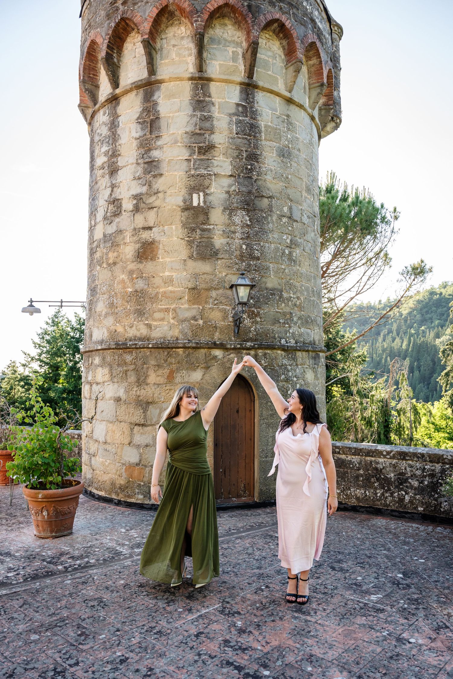Two women in elegant dresses dance joyfully in front of a historic stone tower in a scenic Italian setting.