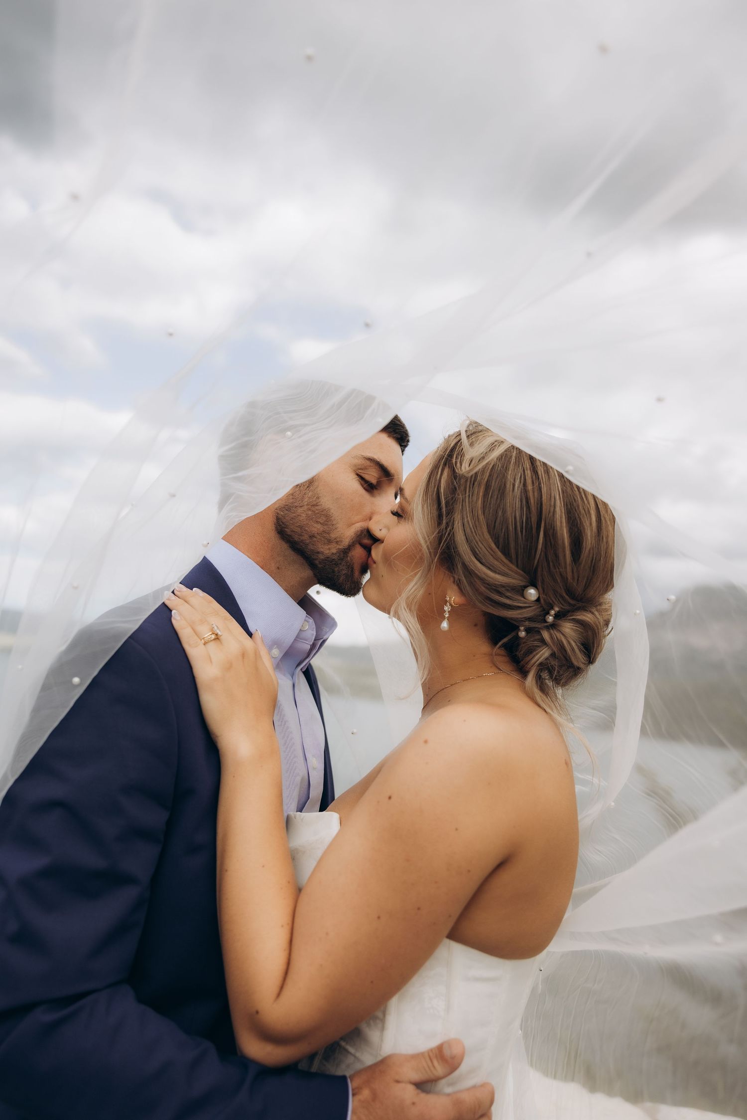 Bride and groom sharing a romantic kiss under a flowing white veil on their wedding day with cloudy sky backdrop.