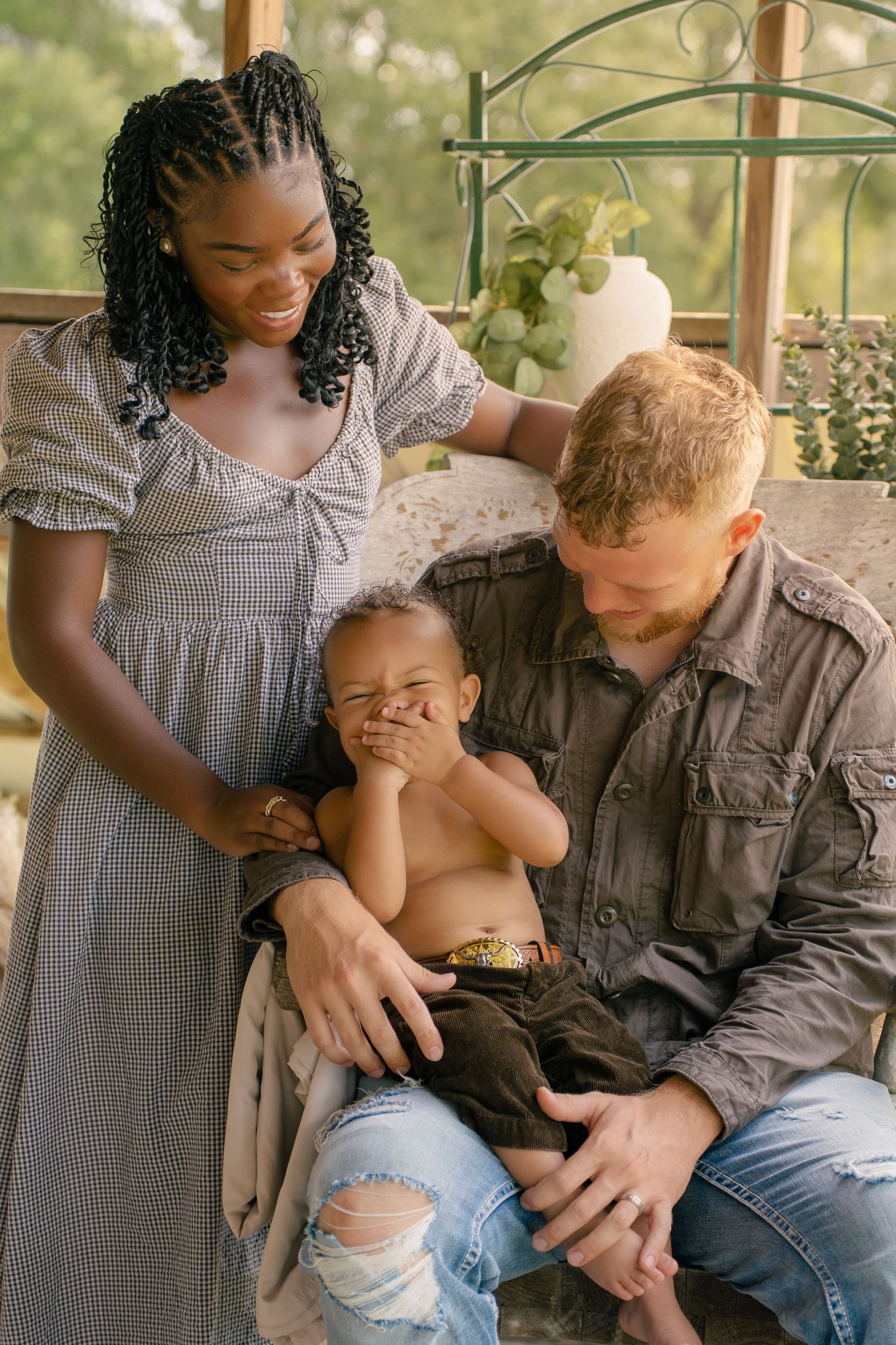 A joyful interracial family with a laughing toddler sitting on dad's lap while mom smiles warmly outdoors.
