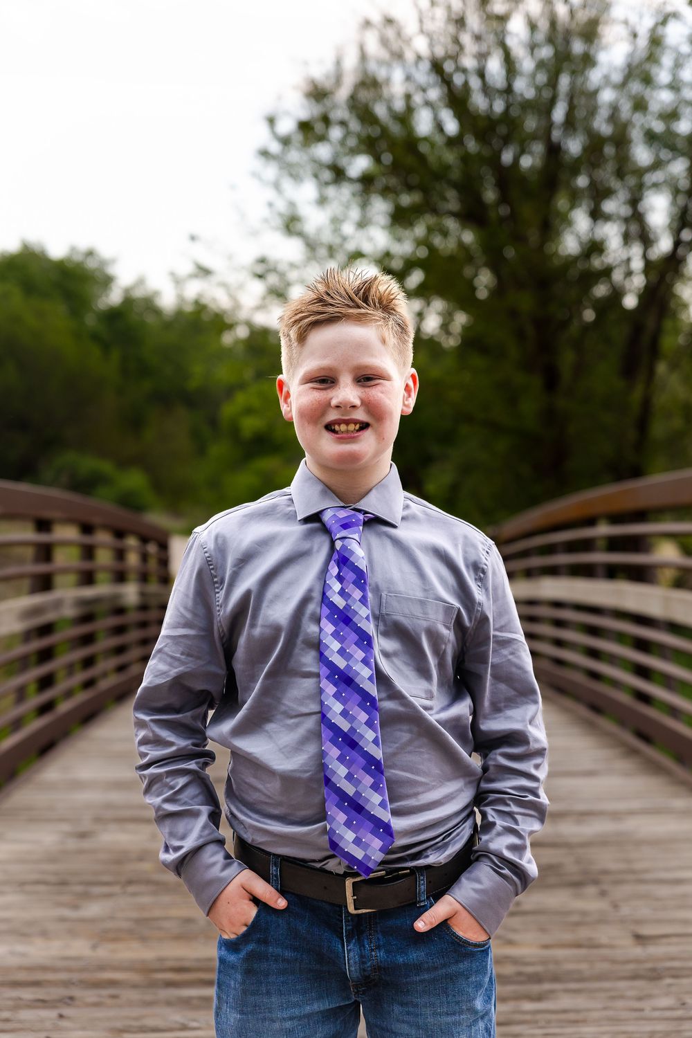 Smiling boy in gray shirt and purple tie stands on wooden bridge with hands in pockets, trees in background.