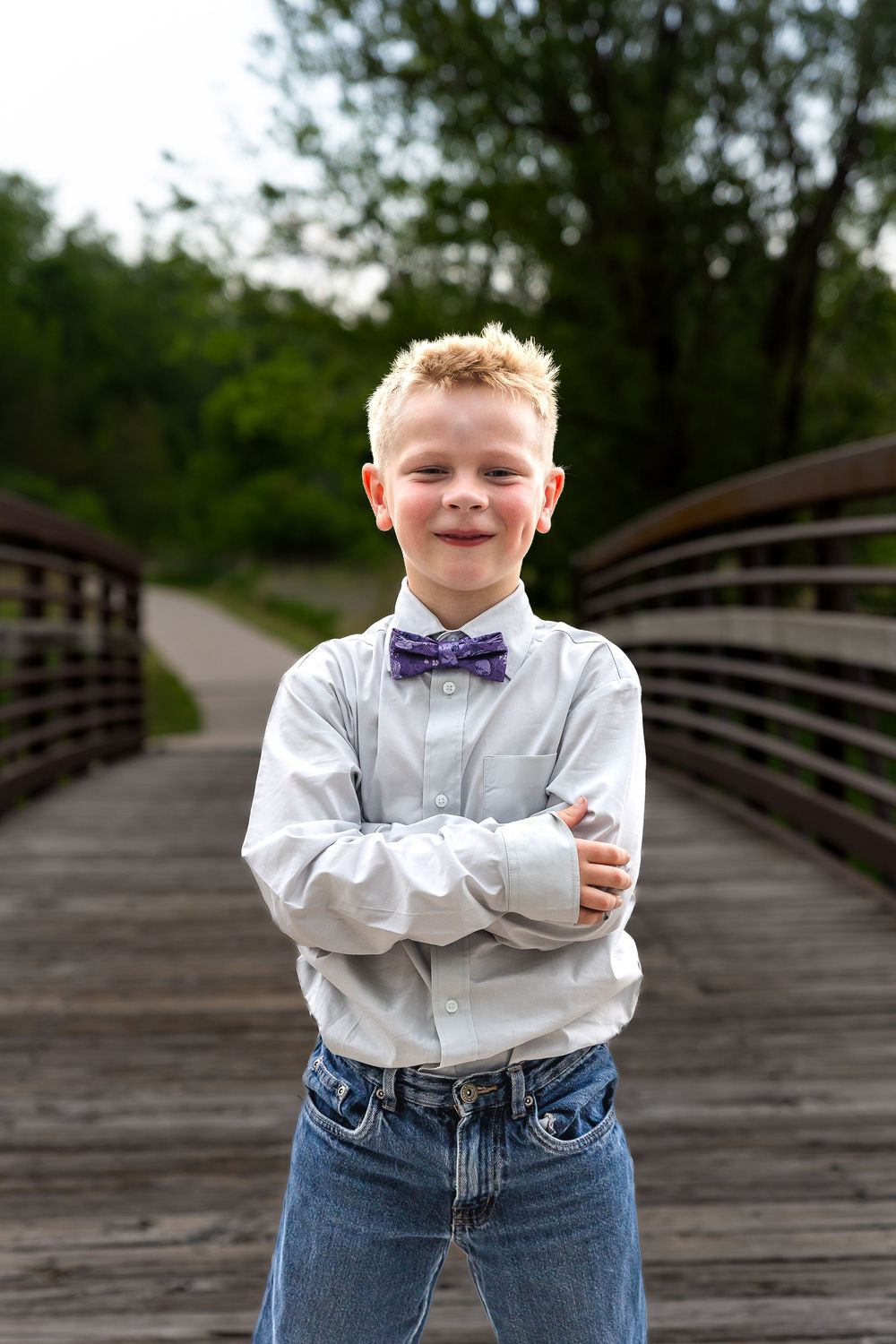 A confident young boy in a white shirt and purple bow tie stands with arms crossed on a wooden bridge surrounded by trees.