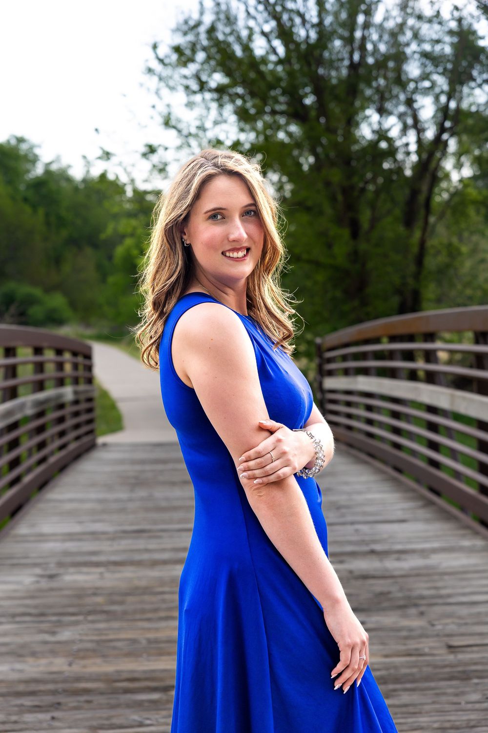 Woman in royal blue dress smiling on wooden bridge surrounded by green trees.