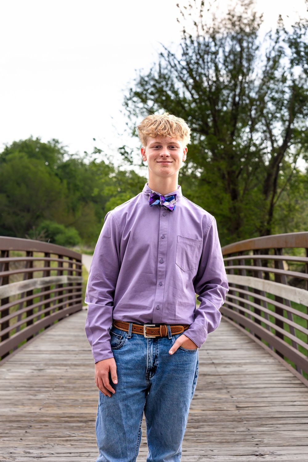 A teenage boy in a purple shirt, bow tie, and jeans smiles on a wooden bridge surrounded by green trees.