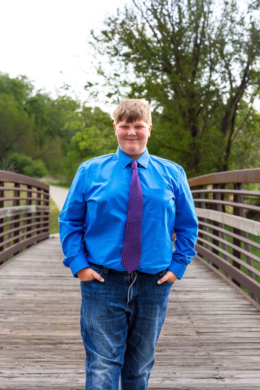A smiling young man in a blue dress shirt and purple tie stands on a wooden bridge surrounded by green trees.