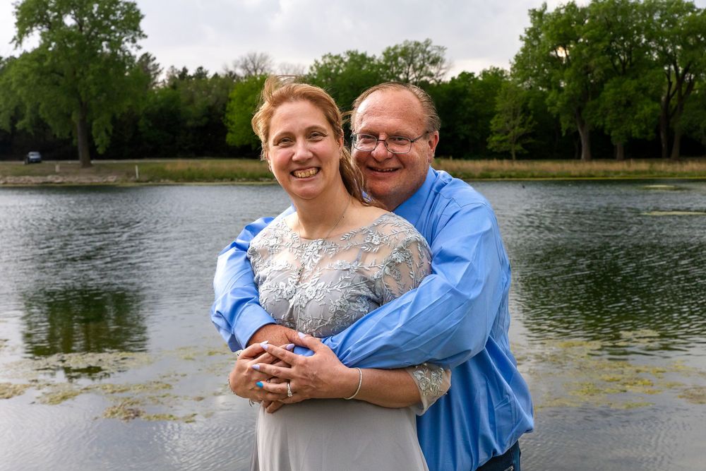 A smiling couple embraces outdoors near a serene lake surrounded by lush green trees on a cloudy day.