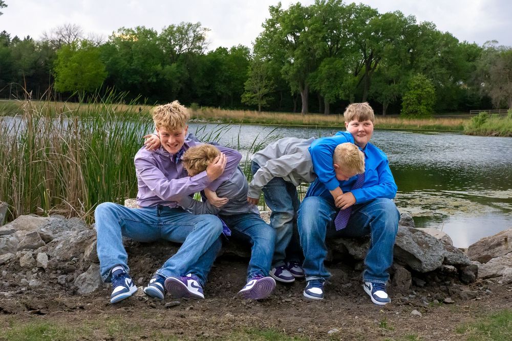 Three kids sit on rocks near a pond with a puppy, surrounded by lush green trees on a cloudy day.