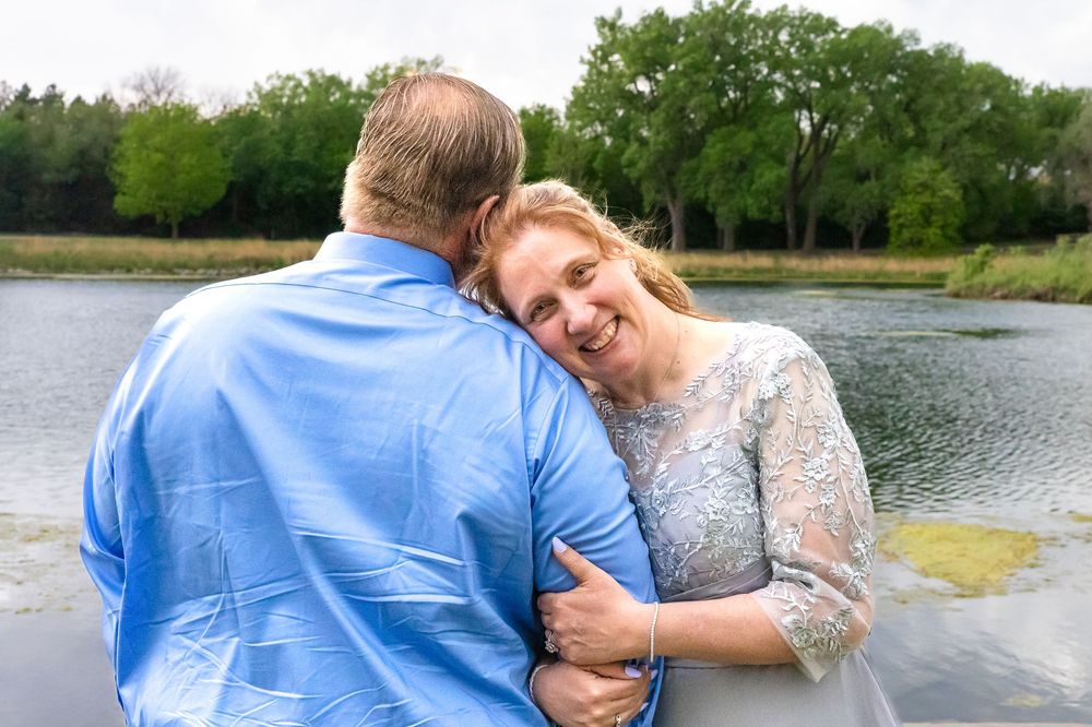 A smiling bride embraces her groom in a blue shirt near a serene lake with lush green trees in the background.