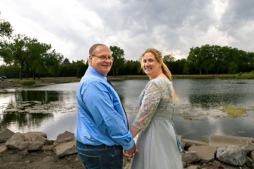 Couple holding hands by a lake, man in blue shirt and woman in lace dress, posing for engagement photos outdoors.