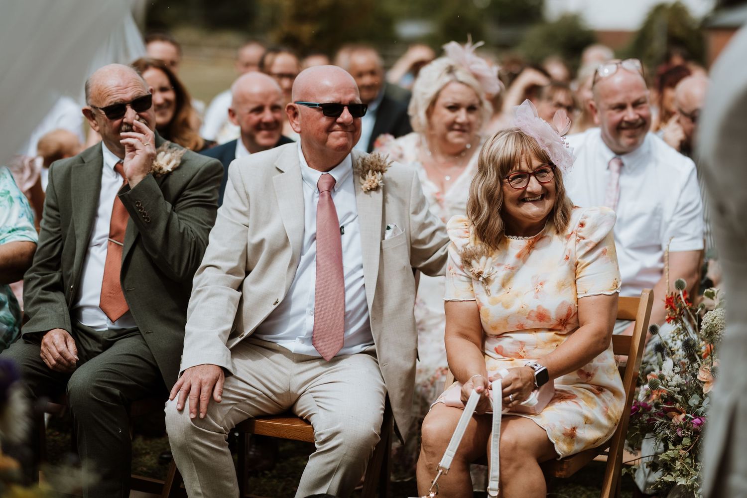 Guests smiling and laughing during an outdoor wedding ceremony, seated on wooden chairs in summer attire.