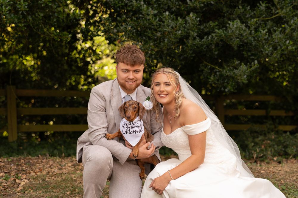 Bride and groom smile with their dachshund wearing a wedding bandana outdoors near green hedges.