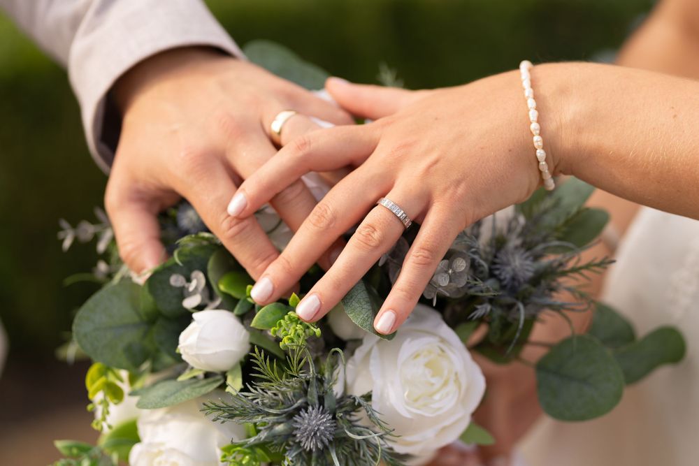 Bride and groom hands with wedding rings resting on a white rose and eucalyptus bridal bouquet.