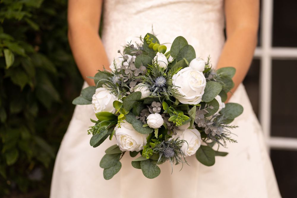 Bride holding a wedding bouquet with white roses, eucalyptus, and blue thistle accents in an elegant arrangement.