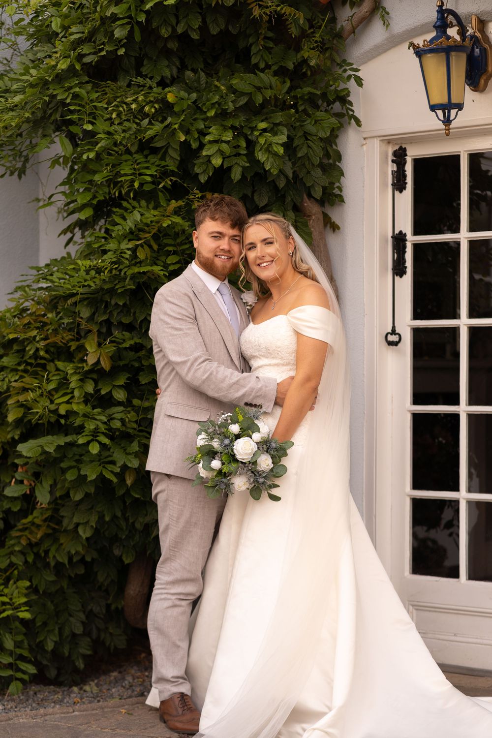 Bride in white off-shoulder gown holding bouquet stands with groom in gray suit outside ivy-covered venue.