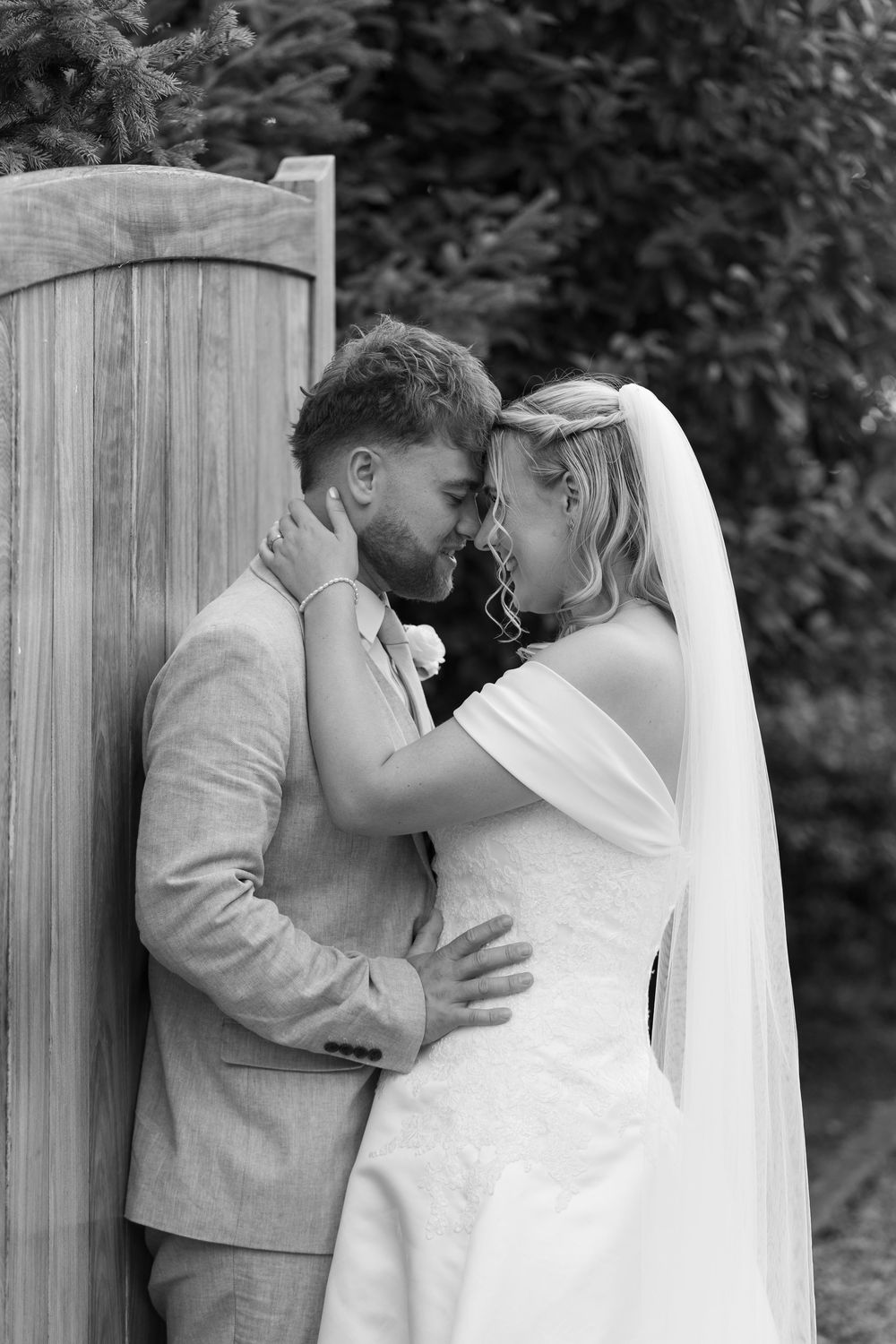 Black and white photo of a bride and groom sharing an intimate moment, foreheads touching, outdoors near a wooden fence.