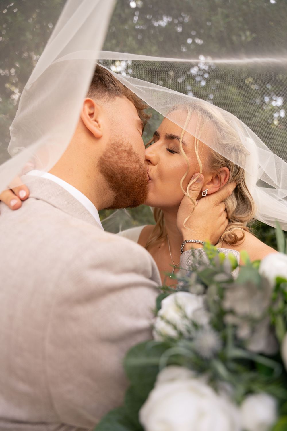 Bride and groom sharing a romantic kiss under a wedding veil, surrounded by white flowers outdoors.