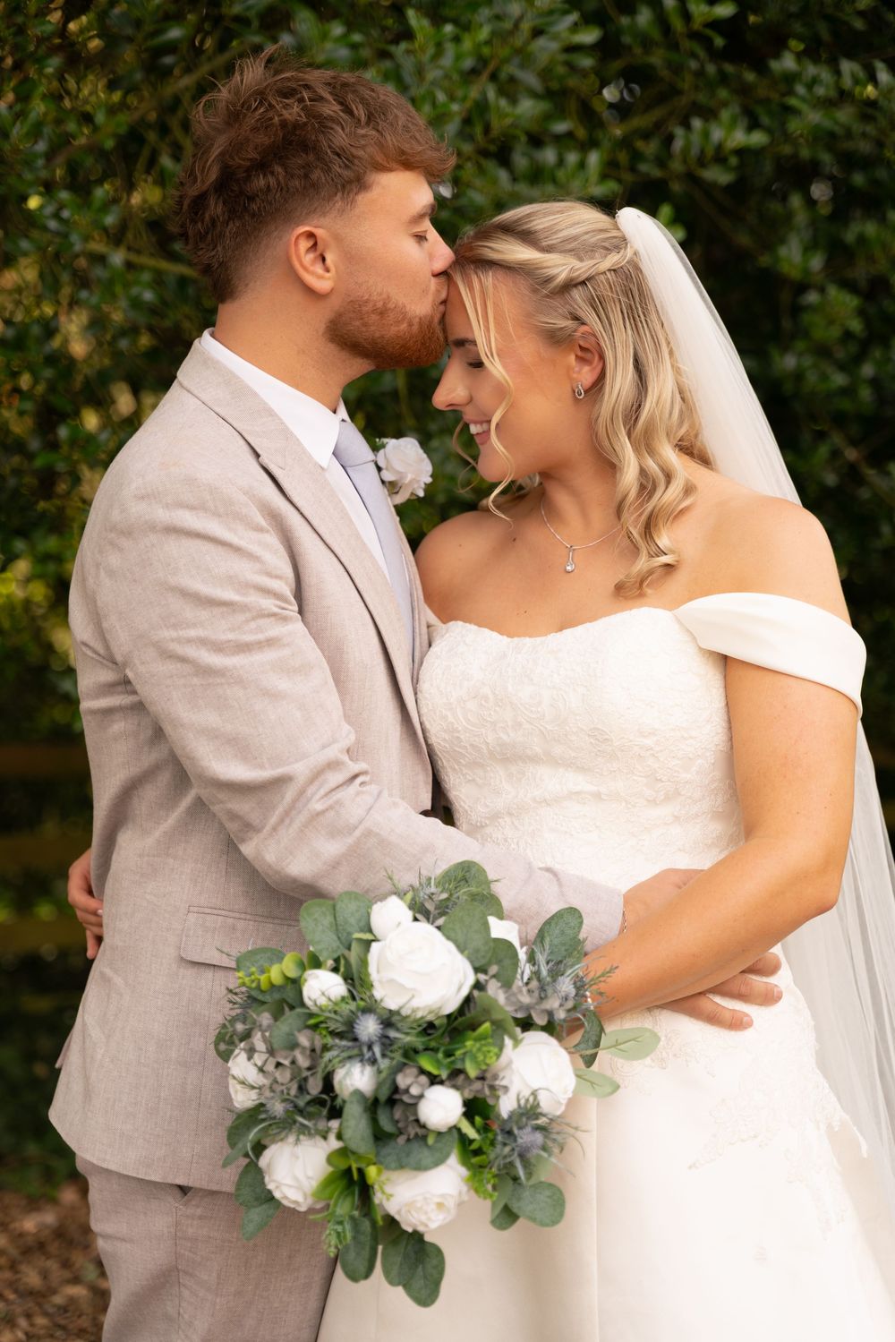 Groom in beige suit kisses bride in white off-shoulder gown holding white floral bouquet outdoors.