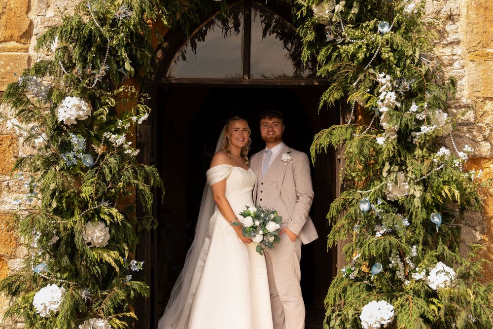 Bride and groom stand beneath floral arch of greenery and white hydrangeas at stone church wedding ceremony.