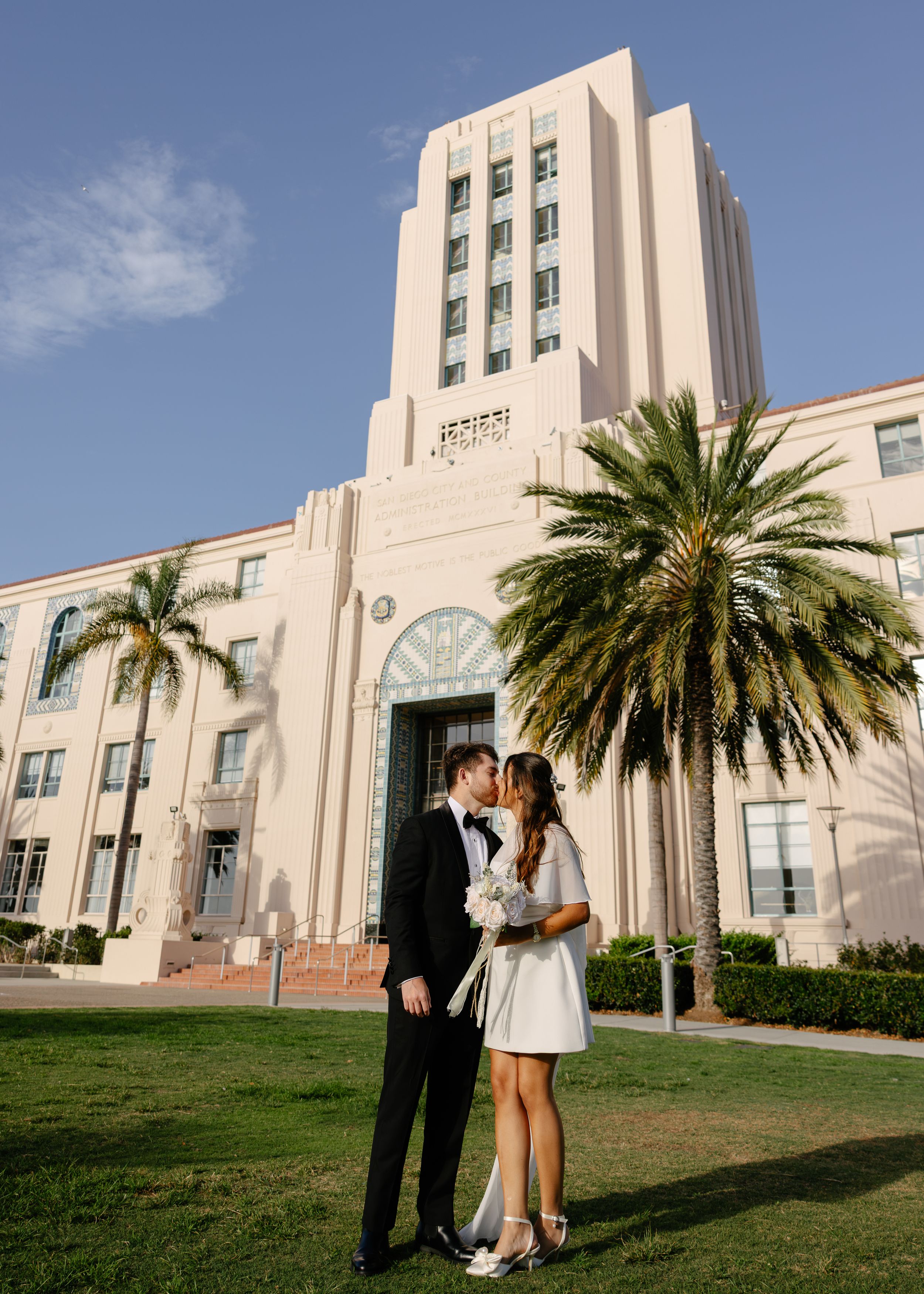 Couple sharing a kiss in wedding attire on a sunny lawn in front of a tall Art Deco building with palm trees.