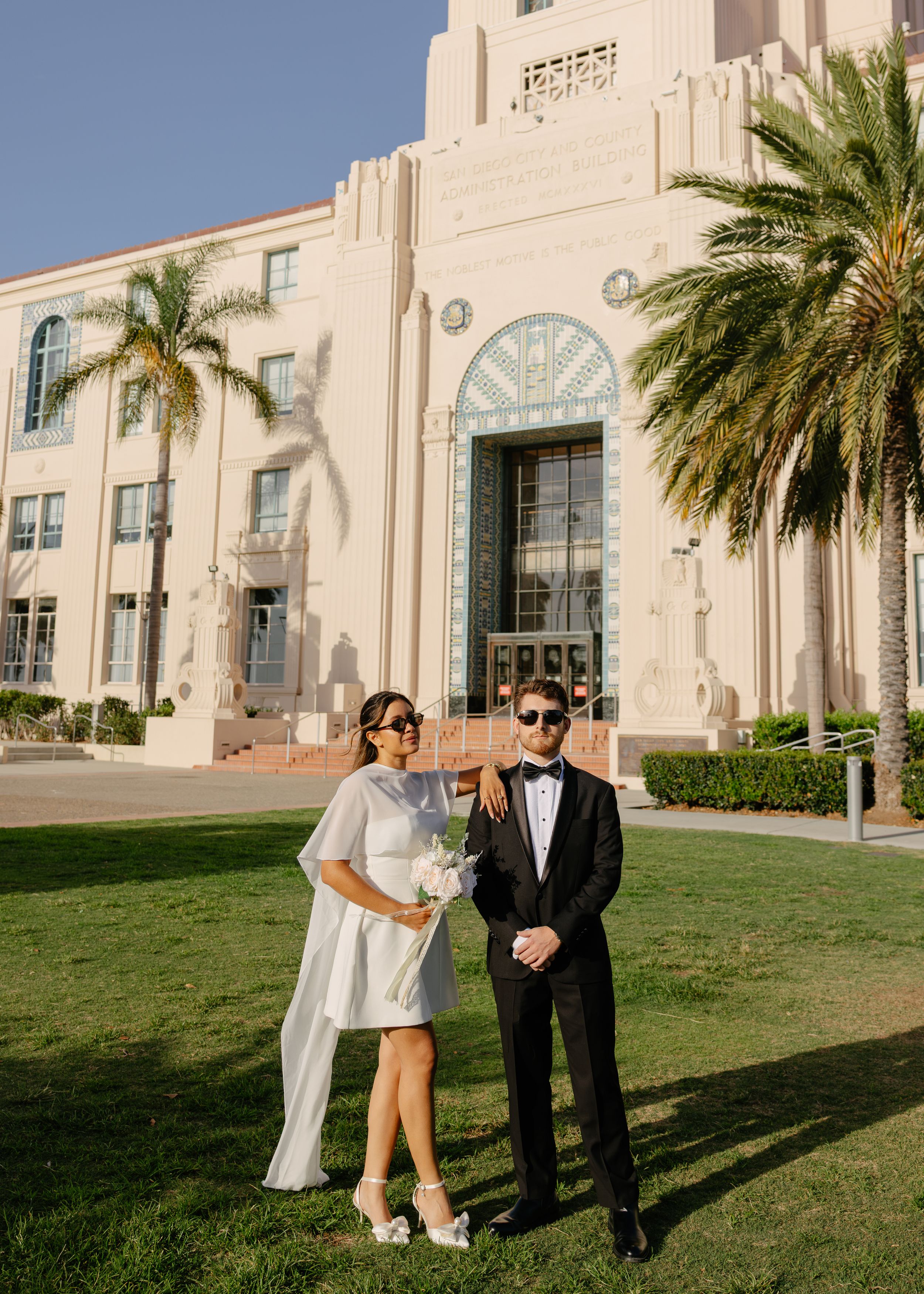 Couple in wedding attire posing on grass before an Art Deco building with palm trees on a sunny day.