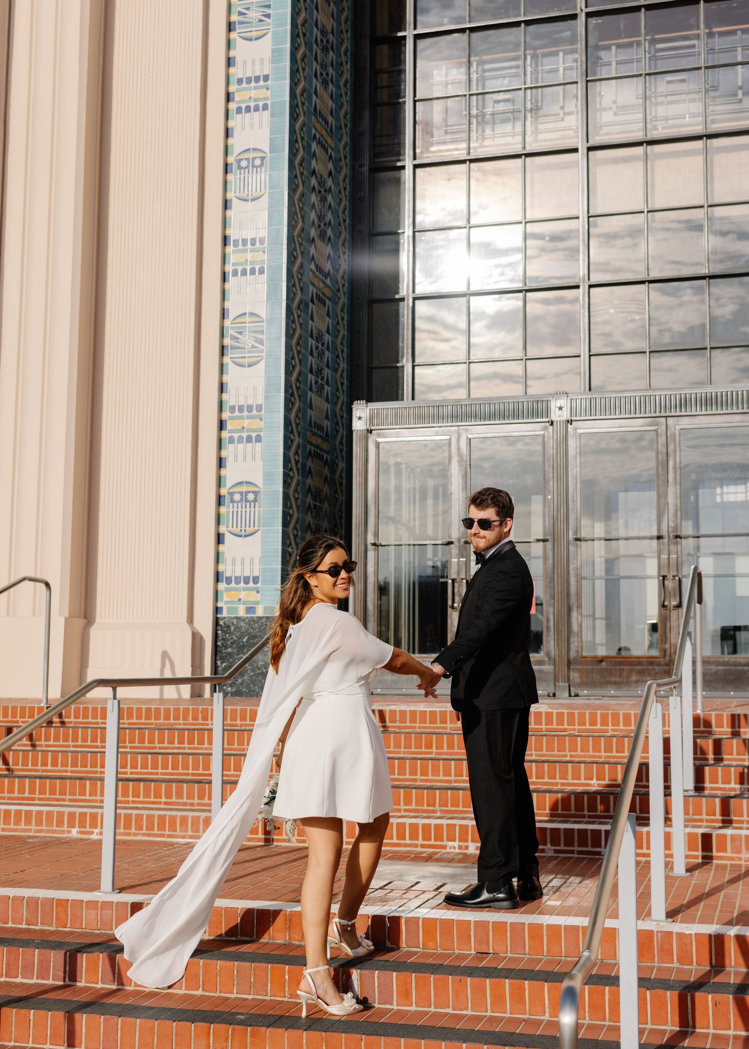 Couple in wedding attire holding hands on brick steps outside a modern building with decorative tiles.