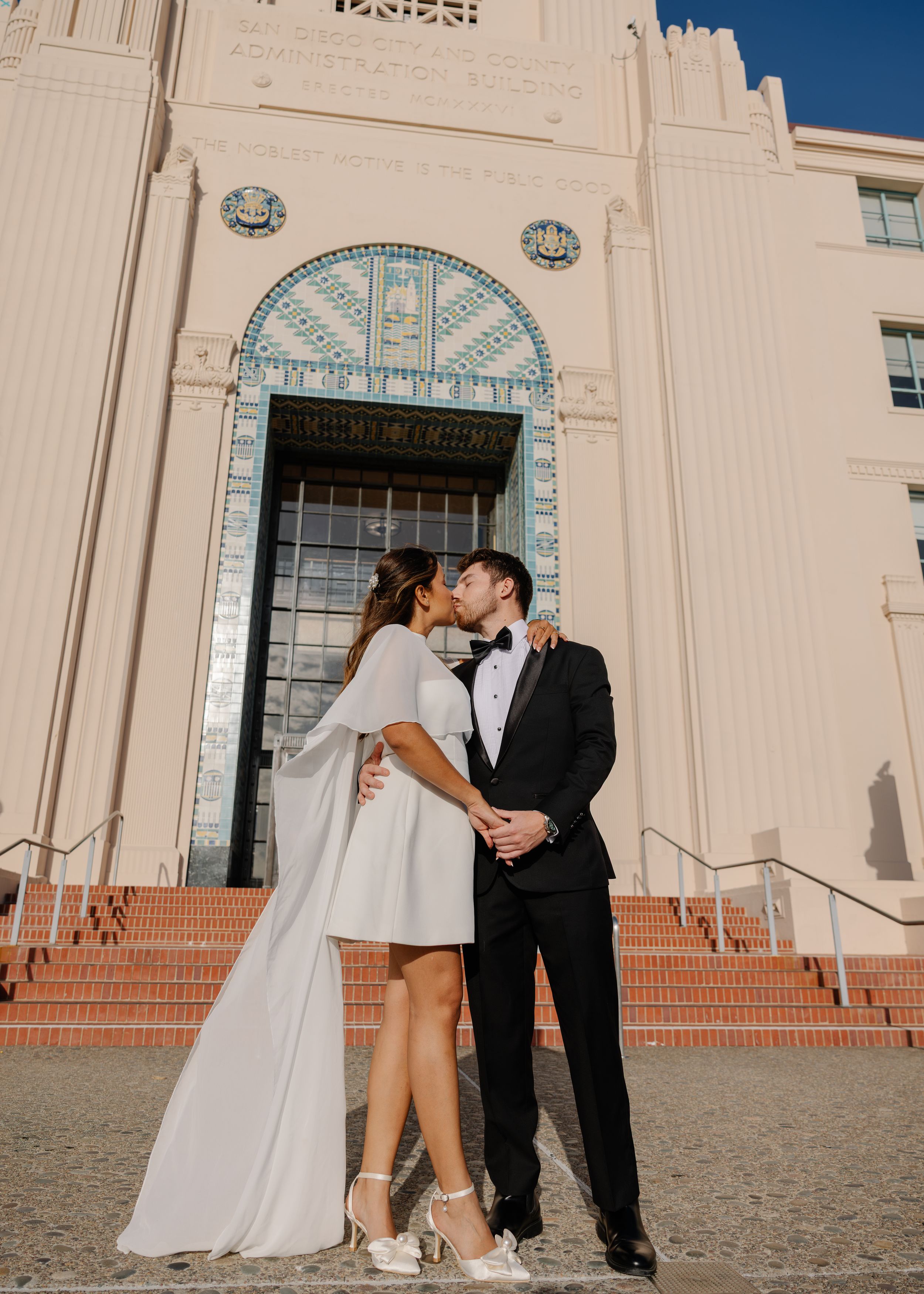 Couple sharing a kiss in wedding attire outside an ornate Art Deco building with tiled archway.