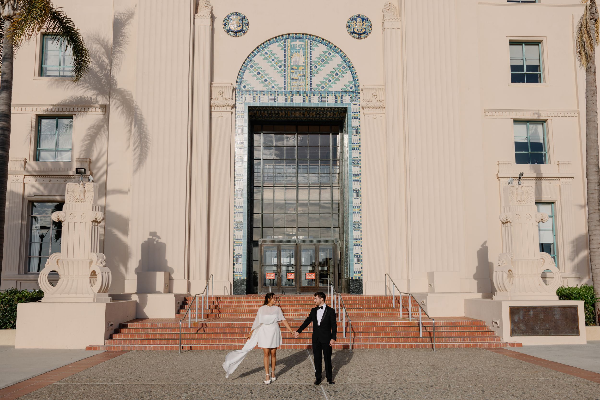 Bride and groom walk hand in hand on red steps outside an elegant Art Deco building on their wedding day.