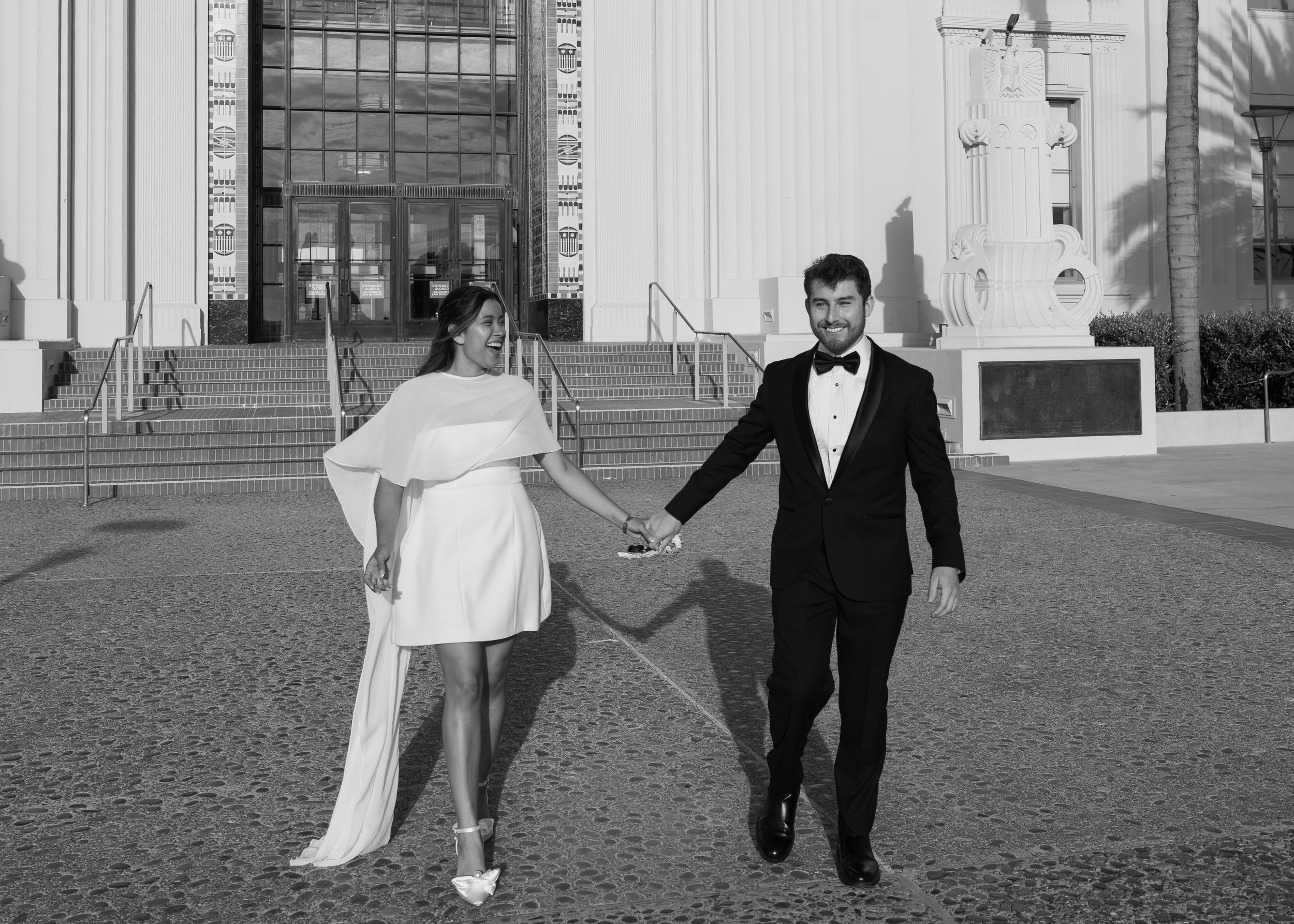 Black and white photo of newlyweds holding hands walking outside a grand building after their wedding.