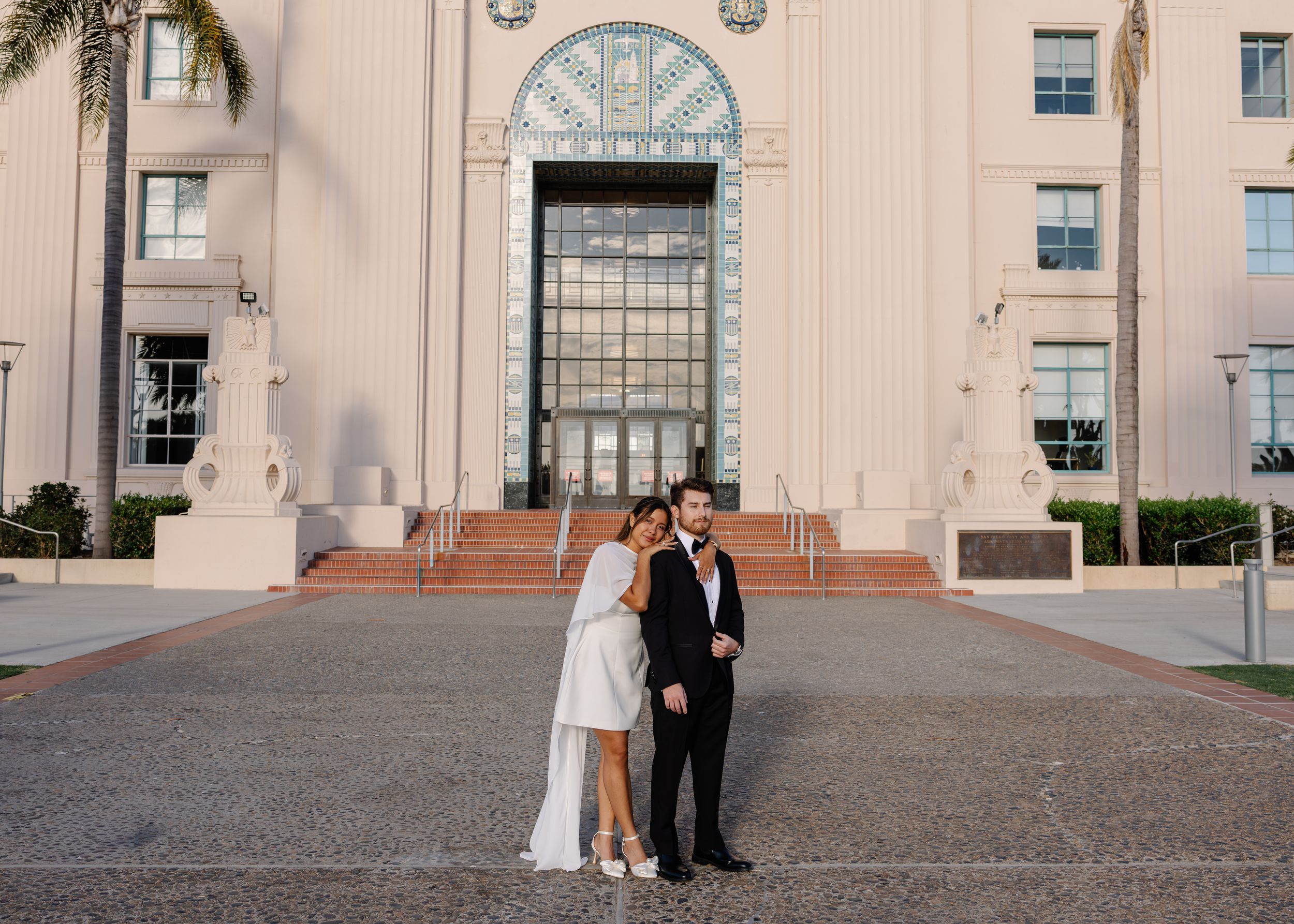 Couple poses in wedding attire before a grand white building entrance on their special day.