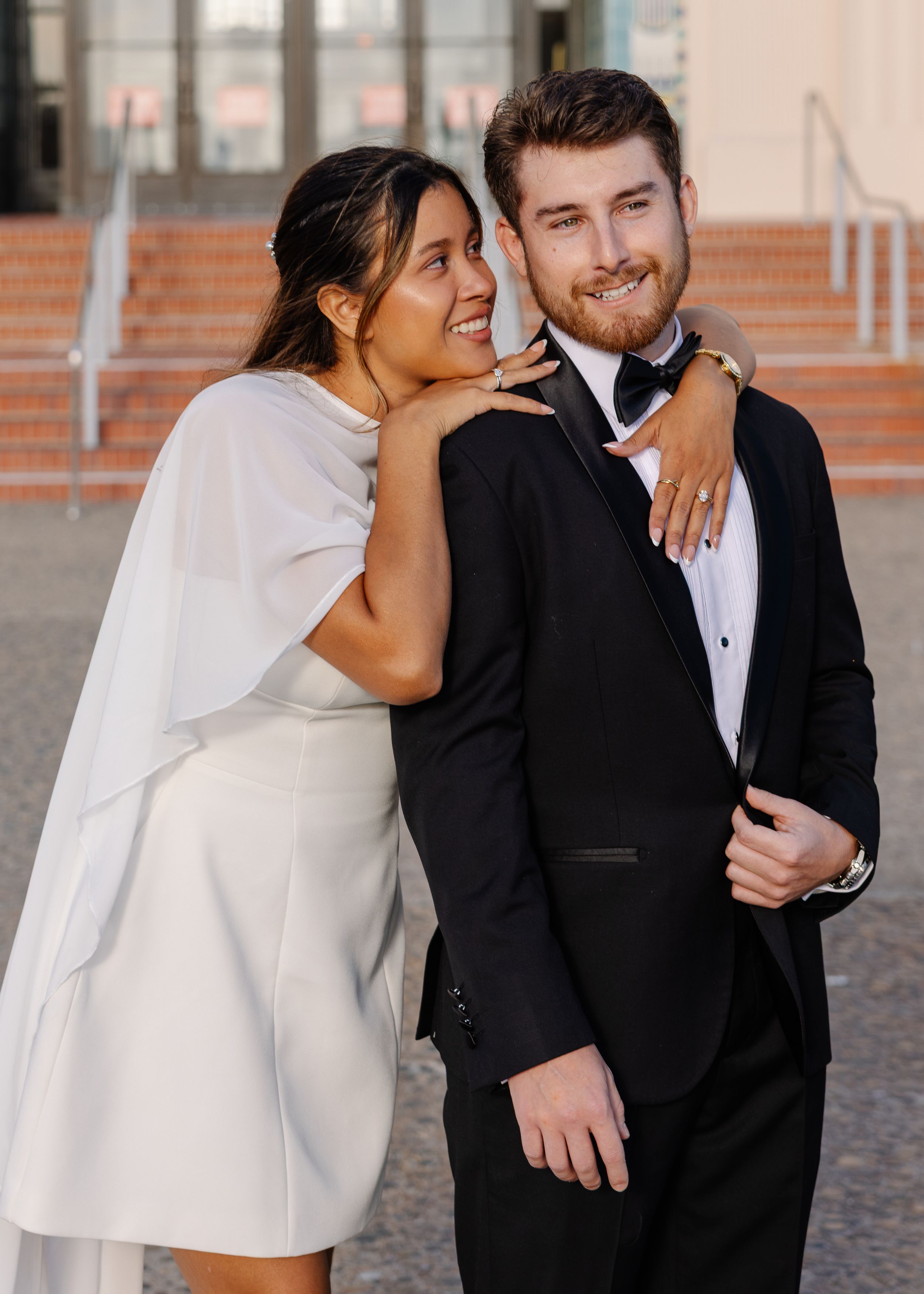A couple in wedding attire poses outdoors, woman in white mini dress embracing man in black tuxedo.