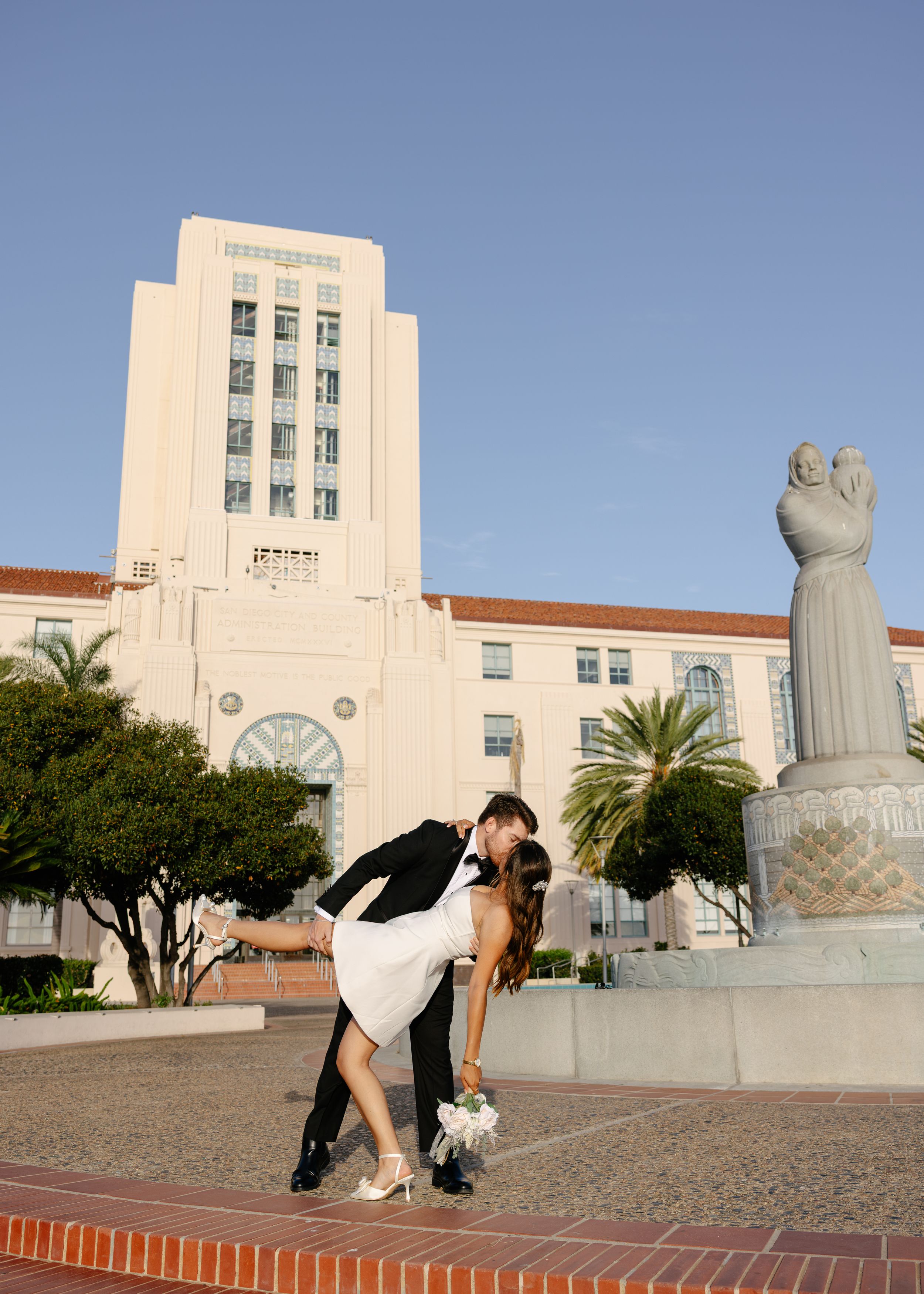 Bride and groom share a romantic dip kiss in front of a grand Art Deco building with a statue fountain.
