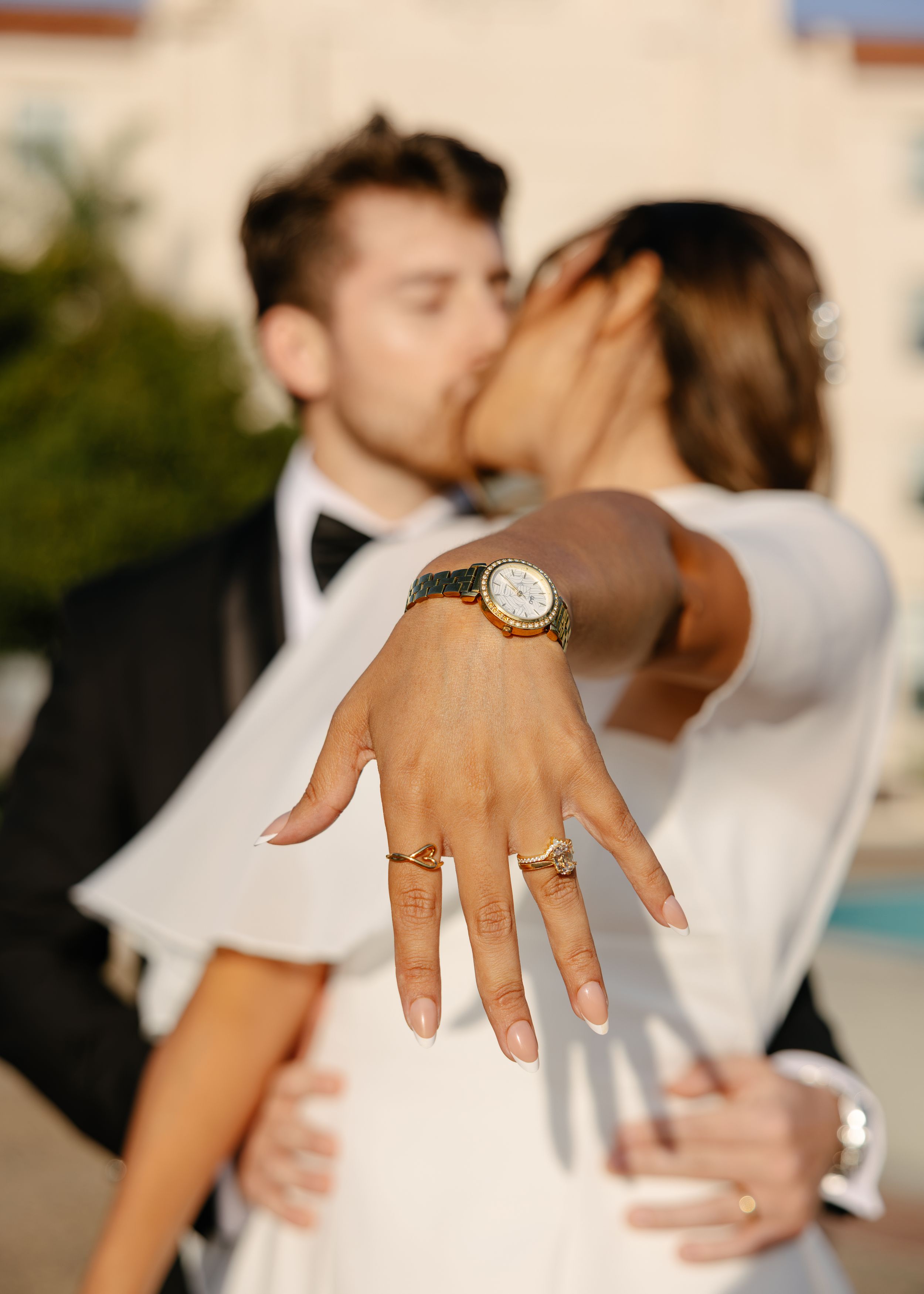 Bride shows off wedding ring and gold watch while kissing groom in tuxedo outdoors.