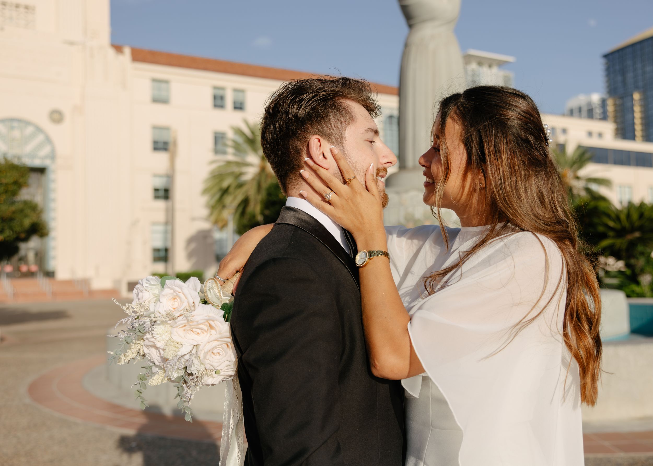A bride and groom share a romantic moment outdoors, with the bride holding a white bouquet and cradling the grooms face.