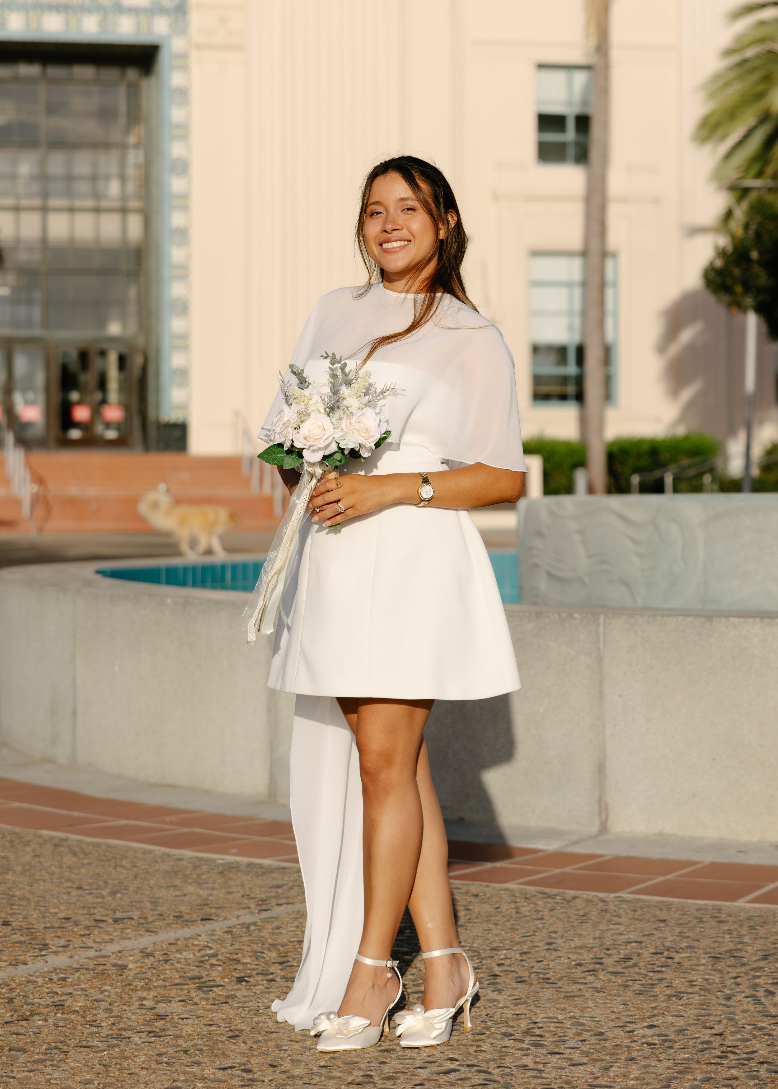 Smiling bride in white mini dress and cape holding white roses bouquet, standing outdoors near fountain.