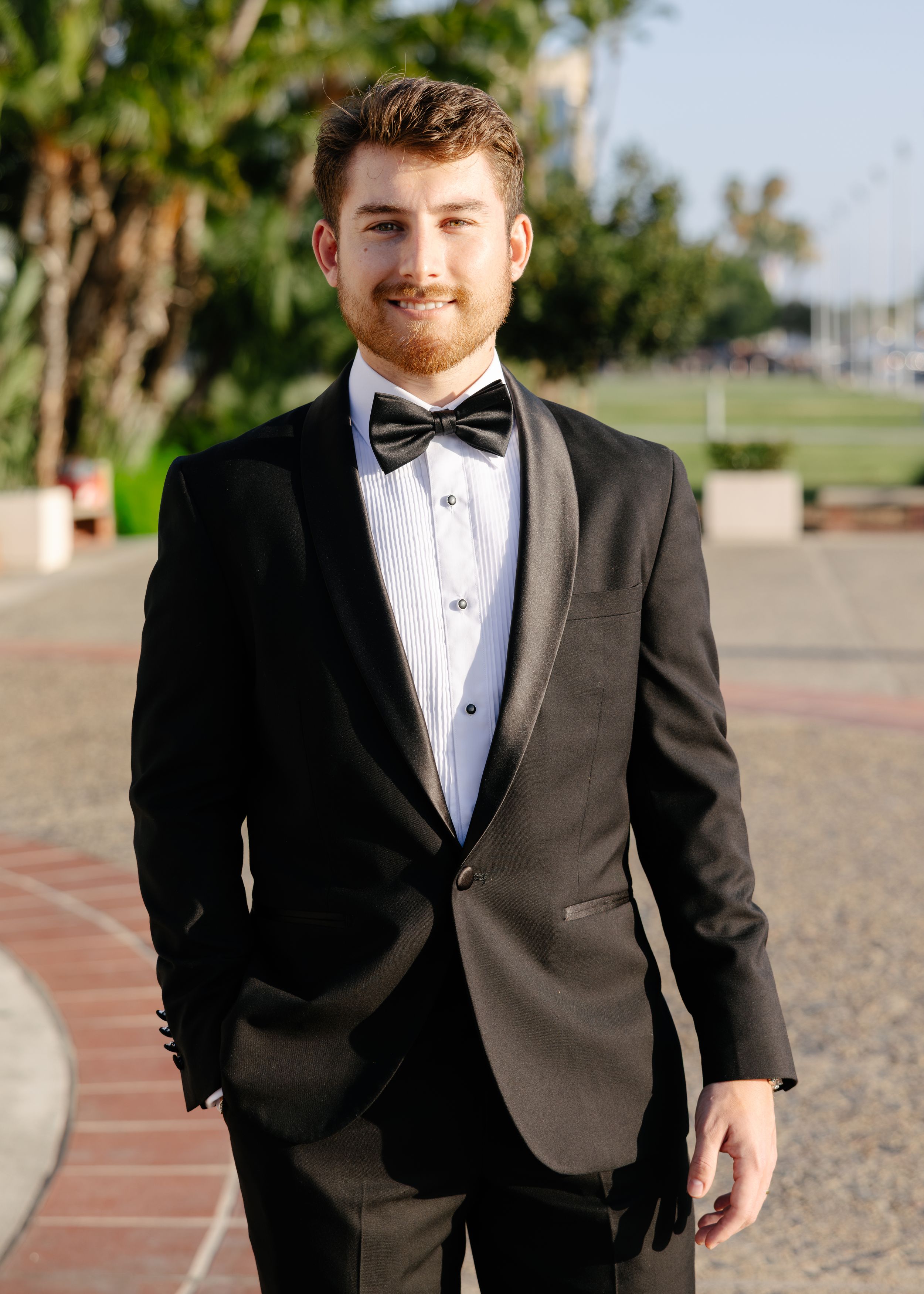 Young man smiling in black tuxedo with bow tie outdoors on brick walkway with trees in background.