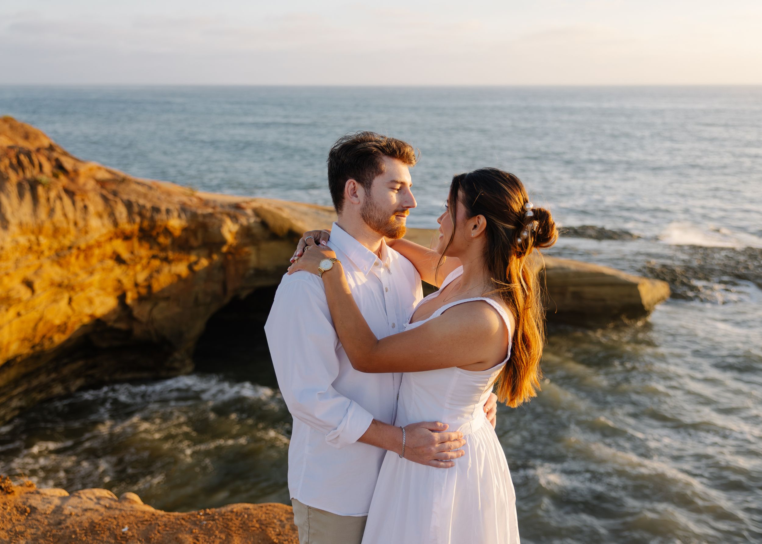 Romantic couple embracing on rocky coastal cliffs with ocean waves at golden hour sunset.