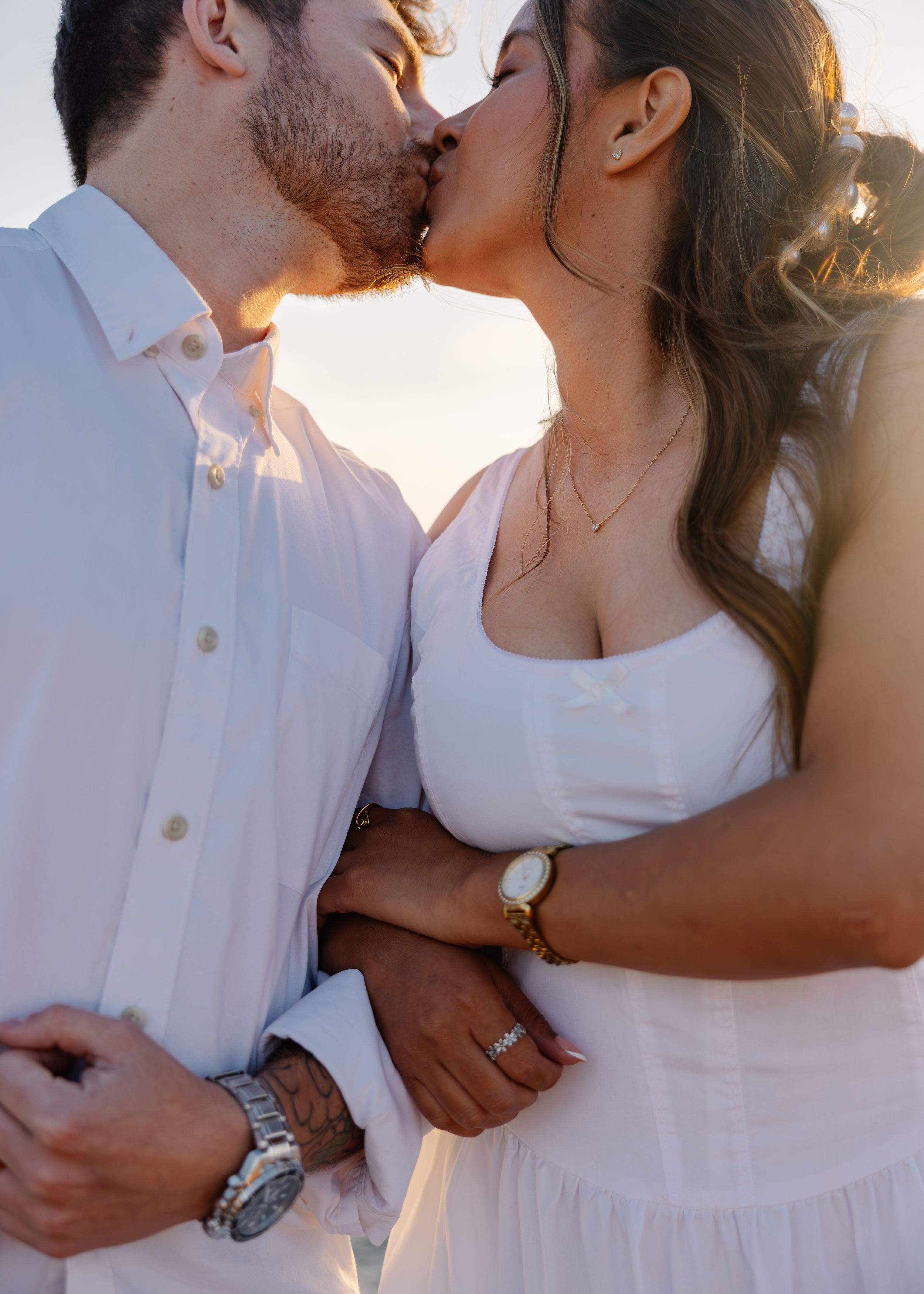 Couple in white outfits sharing a romantic kiss at golden hour, woman wearing a gold watch and ring.