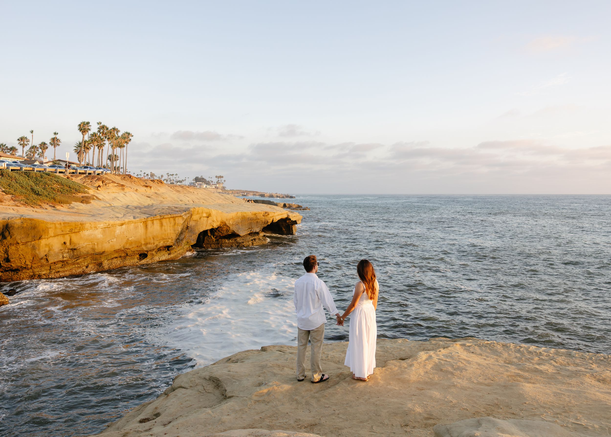 Couple in white holding hands on rocky cliffs overlooking the ocean at sunset in La Jolla, San Diego.