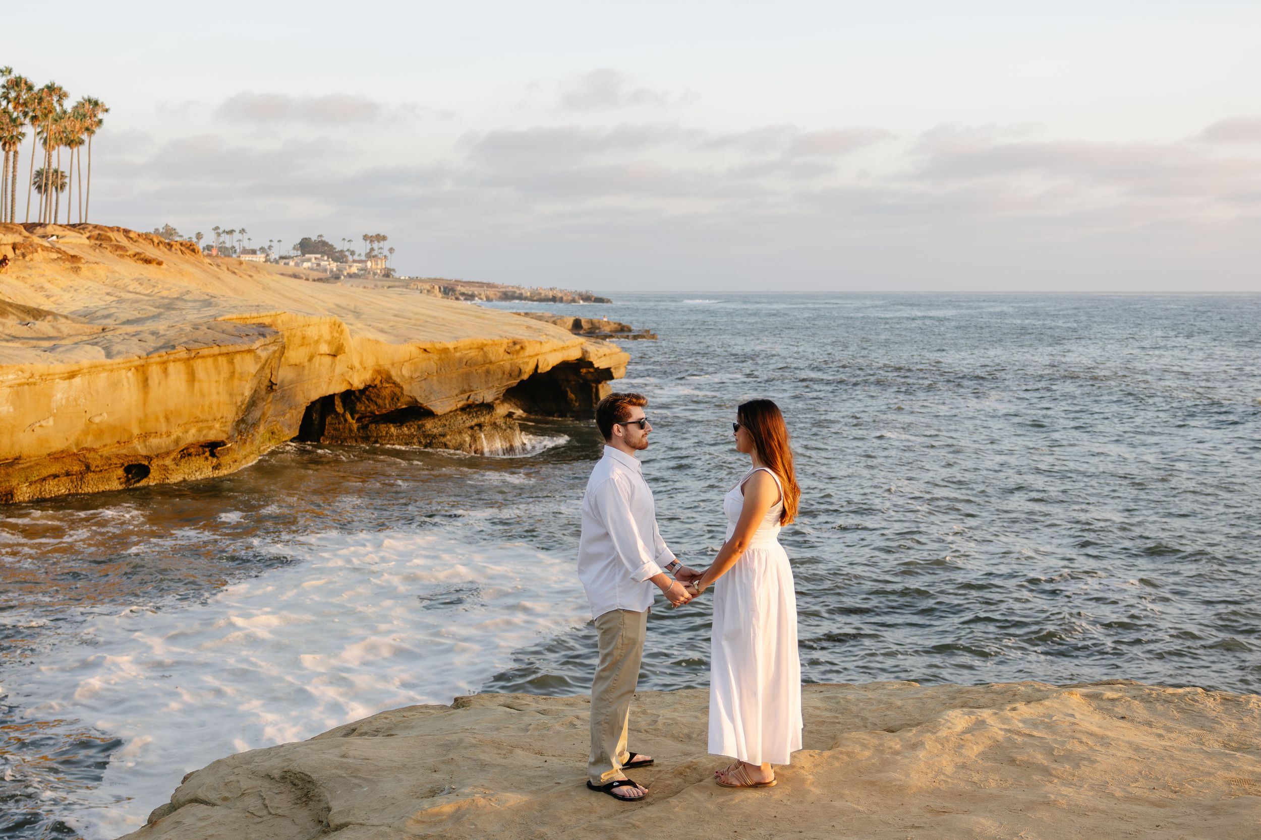 Couple holds hands on rocky coastal cliffs at sunset, waves crashing below, palm trees and ocean in the background.