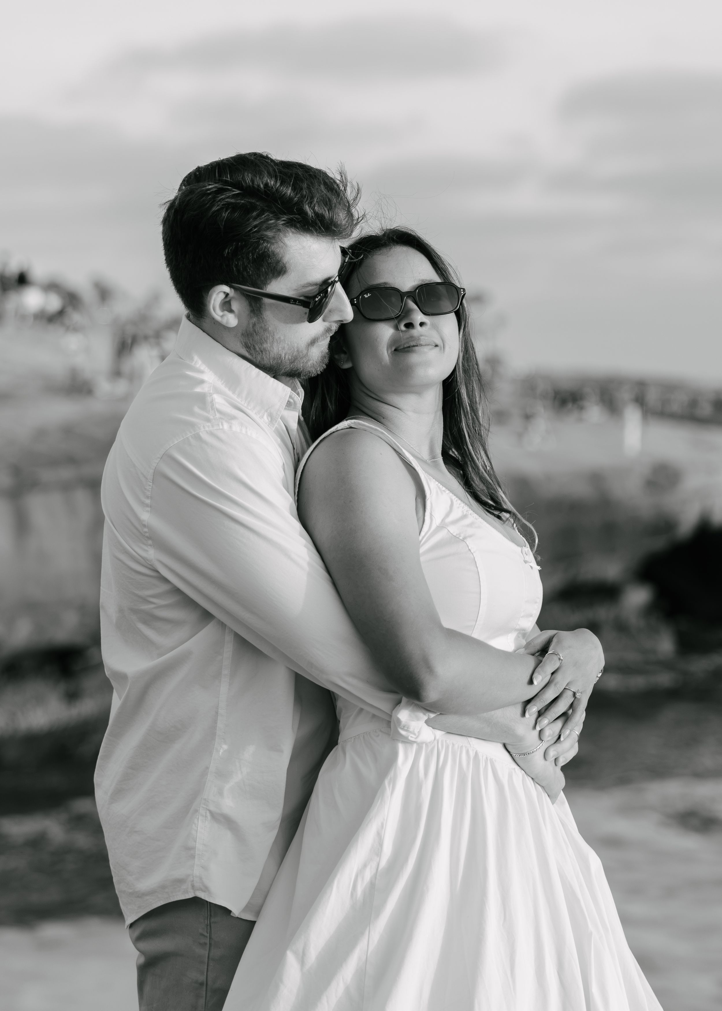 Black and white photo of a romantic couple embracing outdoors, both wearing sunglasses, woman in white dress.