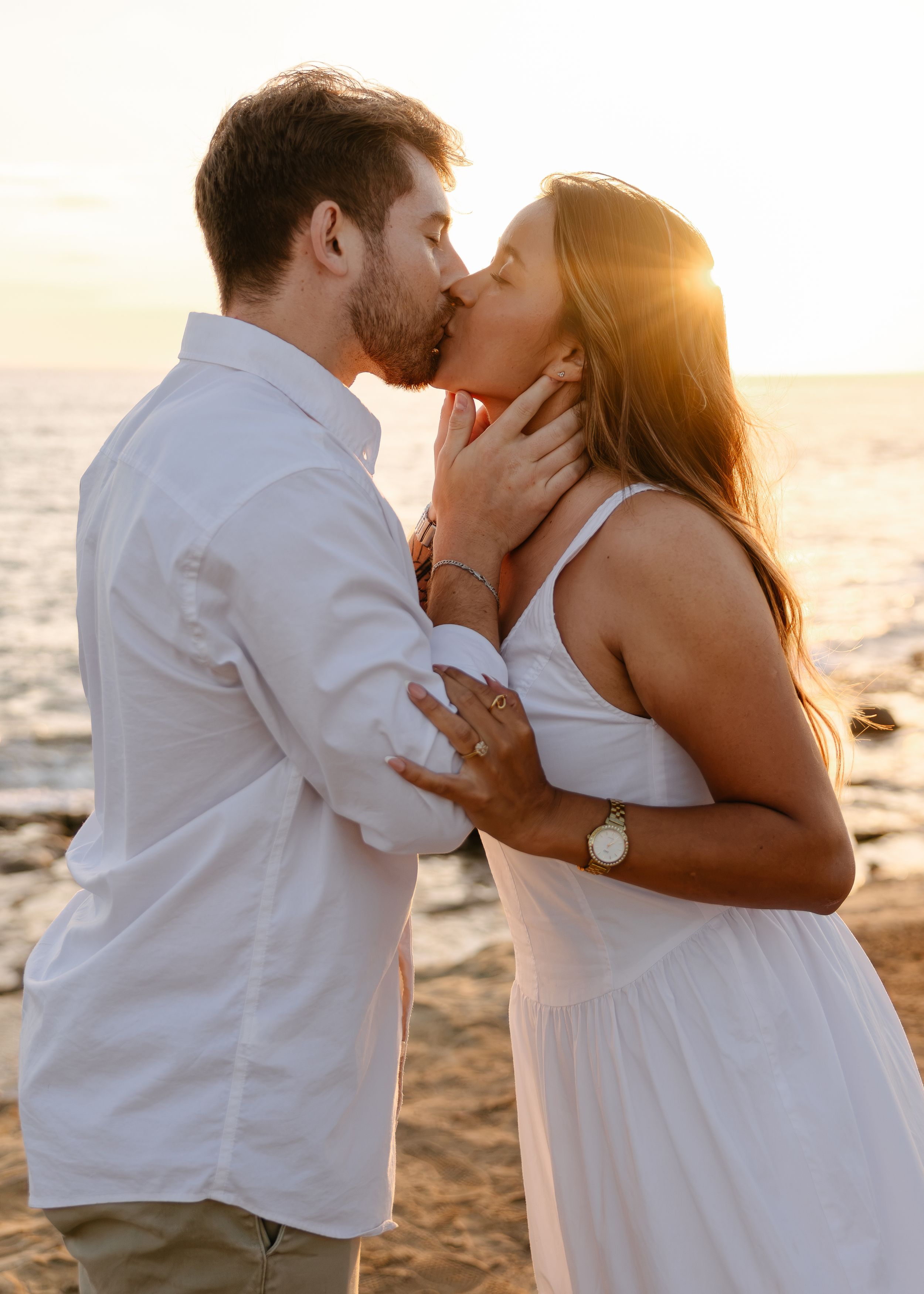 Couple sharing a romantic kiss at sunset on a rocky beach, woman in white dress, golden hour lighting.