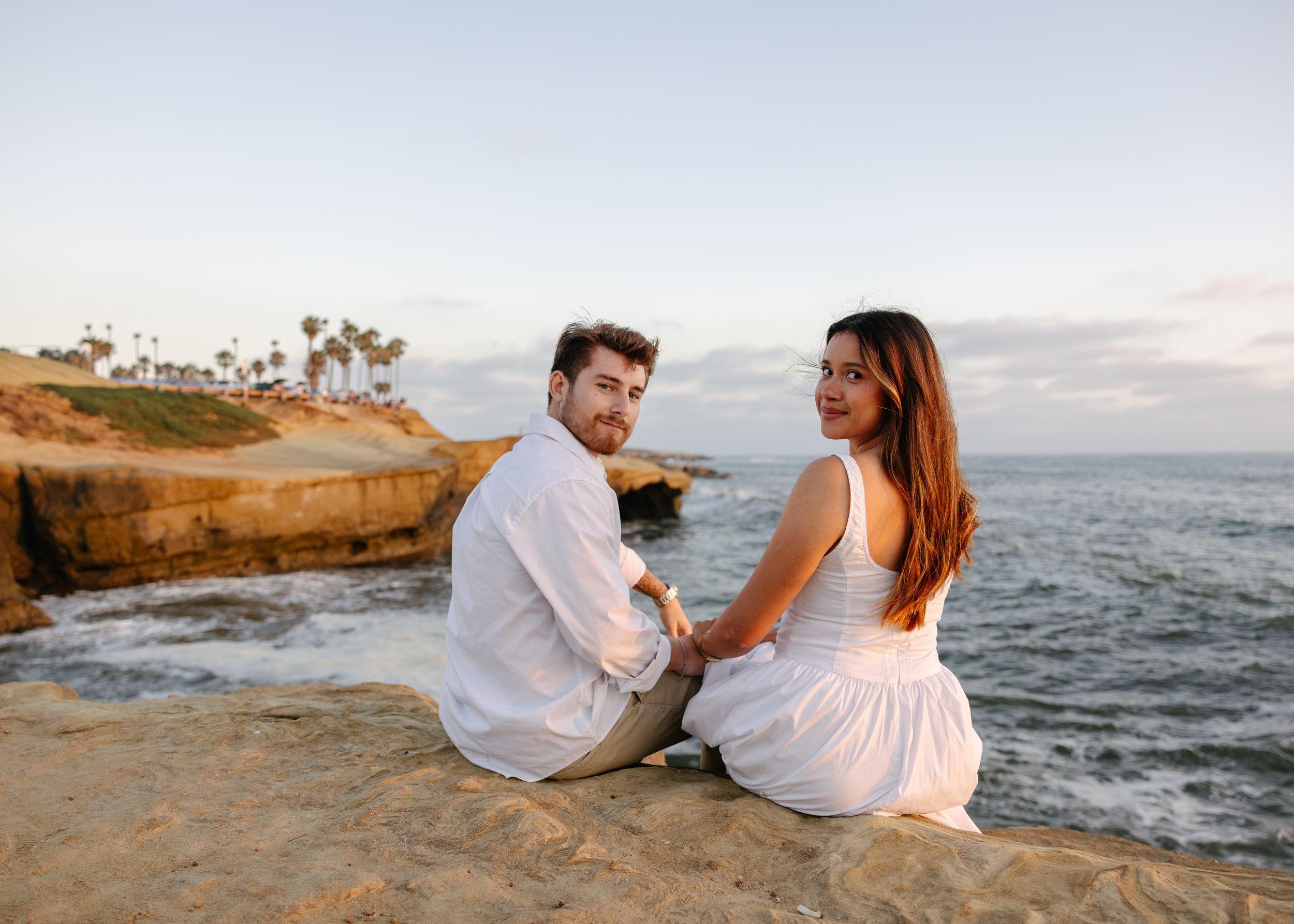 Couple in white outfits sitting on coastal cliffs at sunset, holding hands with ocean and palm trees in background.