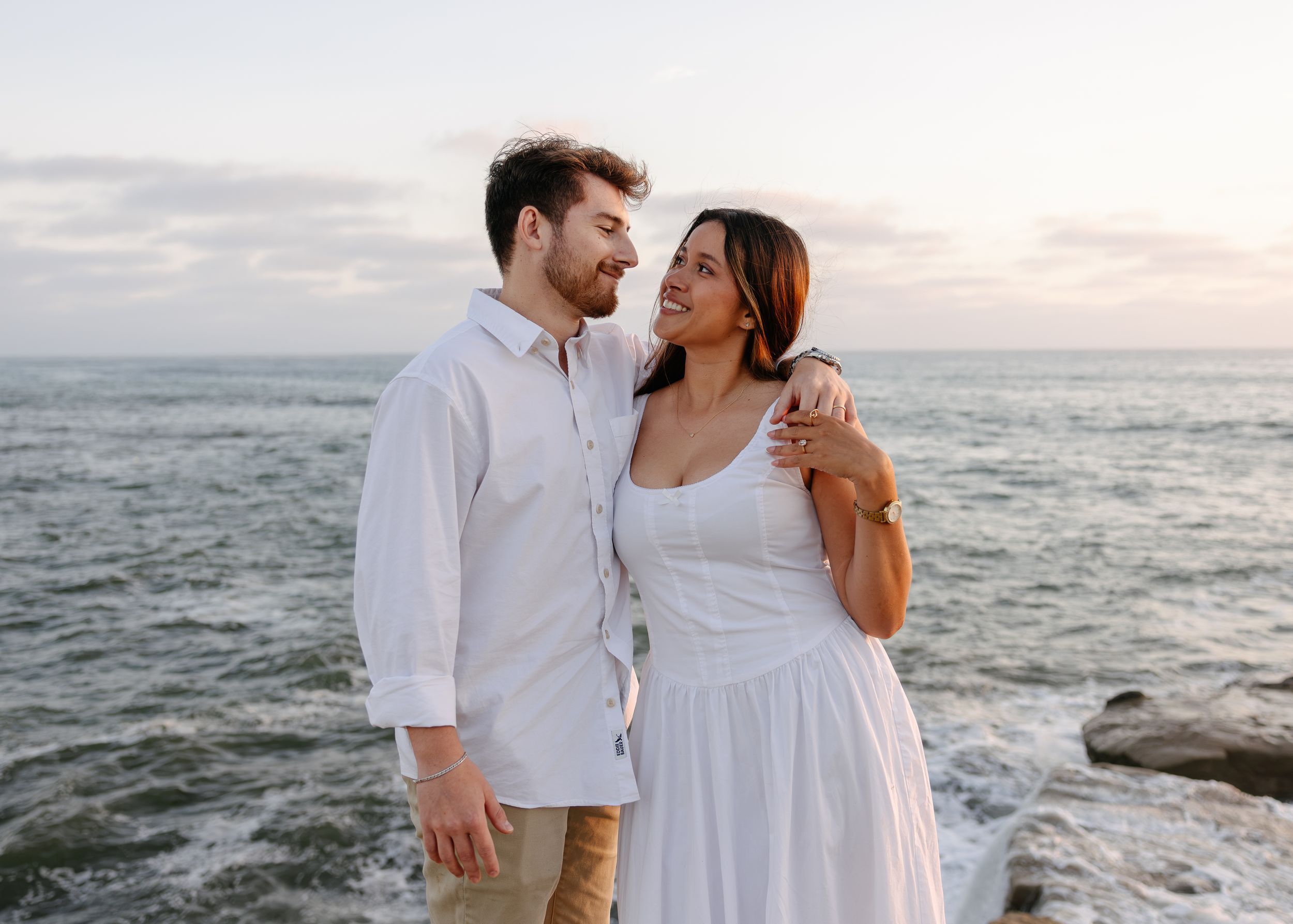 A couple in white outfits smiles at each other on a rocky beach with ocean waves at sunset.