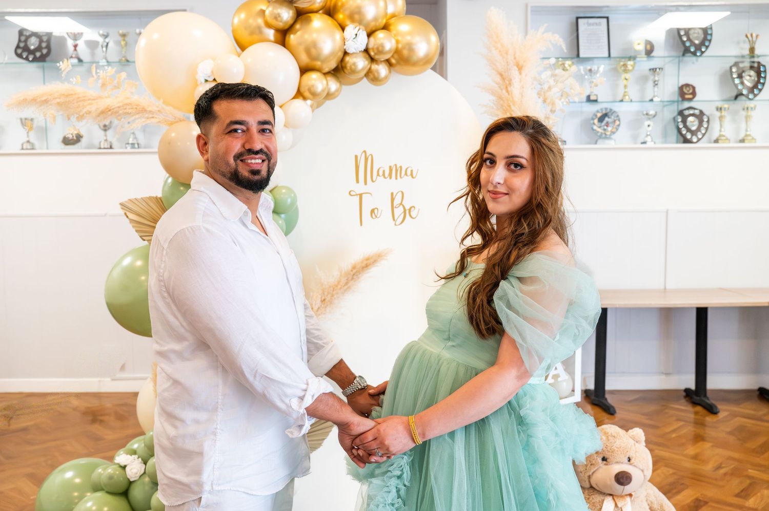 Couple holding hands at a baby shower with a Mama To Be balloon arch backdrop in green and gold.