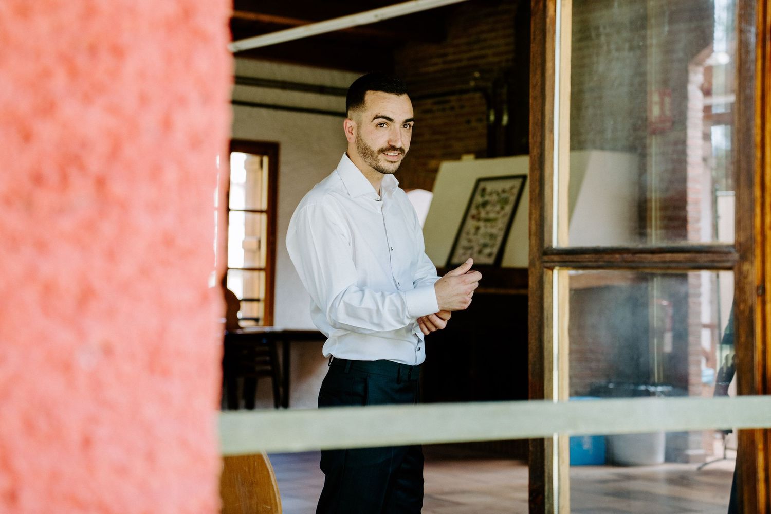 Smiling groom in white shirt adjusts cufflinks while standing in doorway before wedding ceremony.
