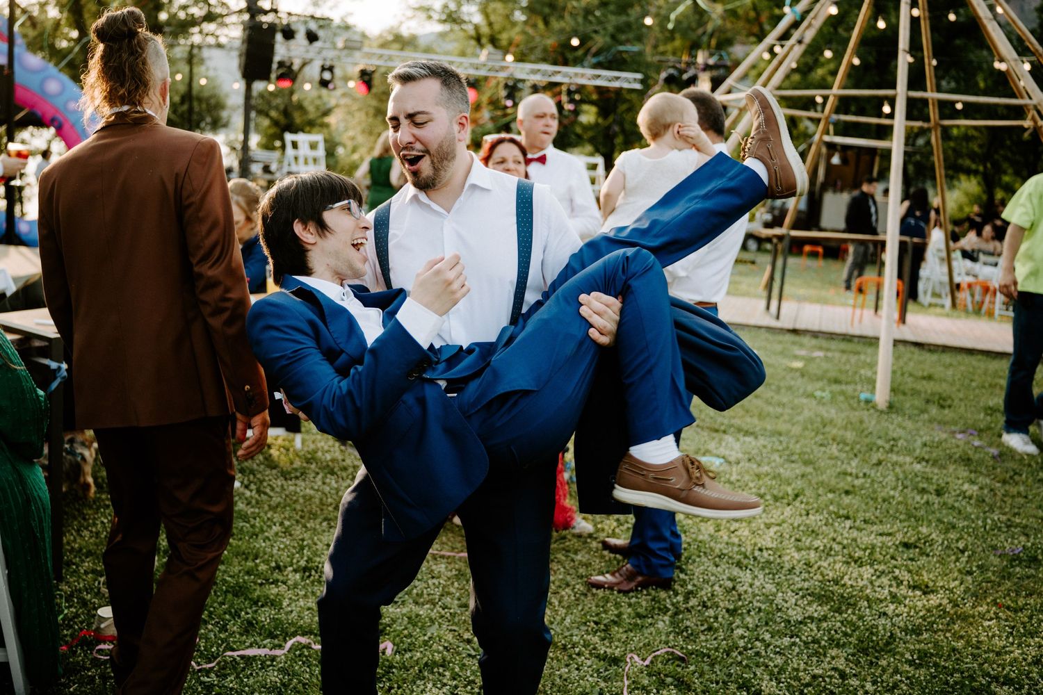 Two men in blue suits playfully dancing and lifting each other at an outdoor wedding reception.