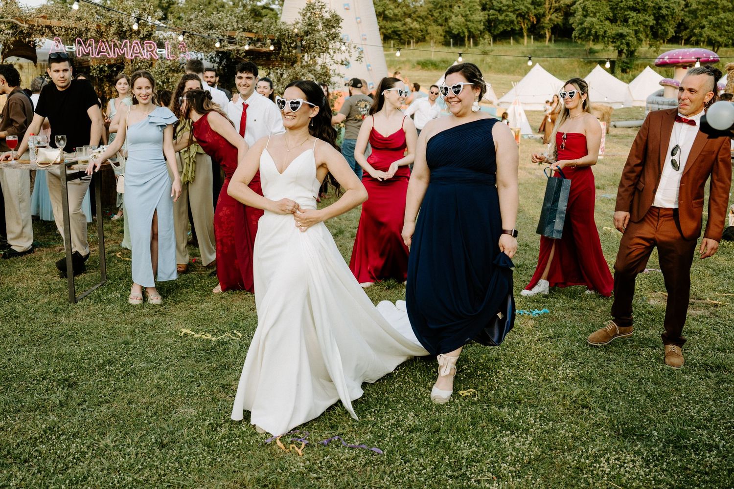Bride in white gown dancing with guests in colorful formal attire at an outdoor wedding reception on green lawn.