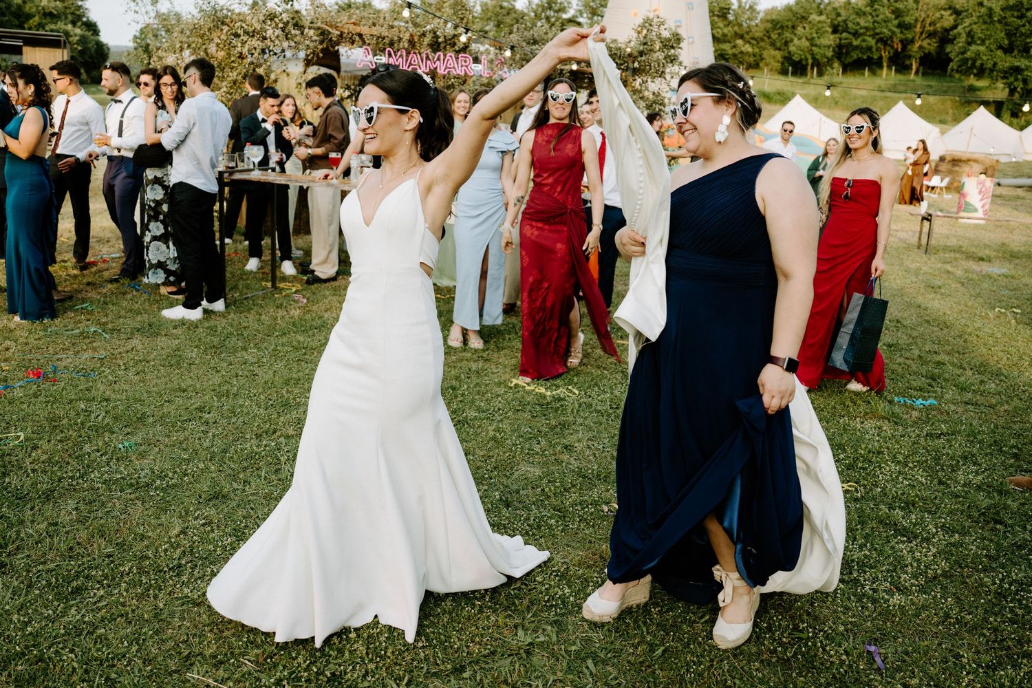 Bride in white gown dancing joyfully with guest in navy dress at outdoor wedding reception.